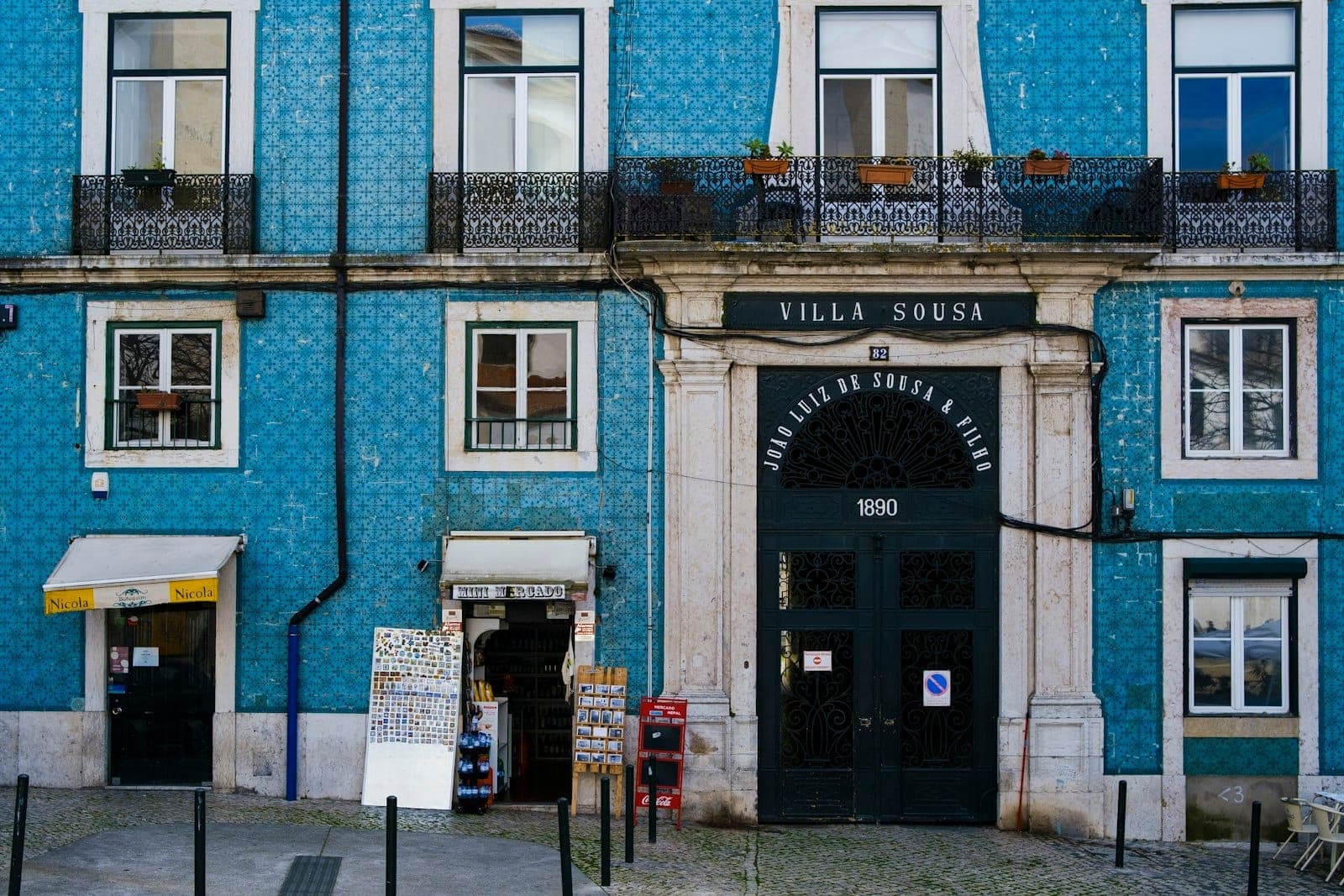 Fachada de um prédio lisboeta tradicional coberto de azulejos azuis, com varandas, uma pequena loja e um arco imponente com a inscrição 'Villa Sousa'.