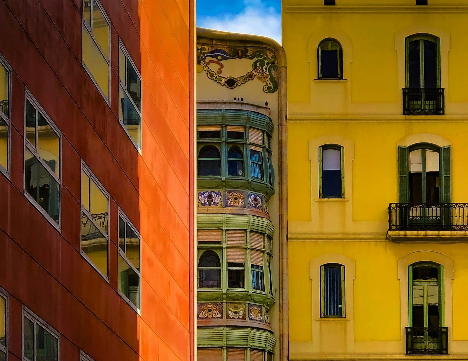 Vista colorida e detalhada dos prédios de Gràcia, com fachadas amarelas e laranjas vibrantes, sacadas ornamentadas e janelas trabalhadas sob um céu azul.