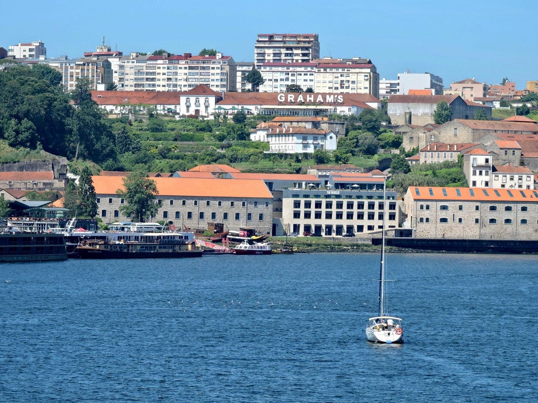 Blick auf Graham's Port Lodge auf einem Hügel über dem Douro in Vila Nova de Gaia, mit traditionellen Gebäuden und einem Segelboot auf dem Wasser.
