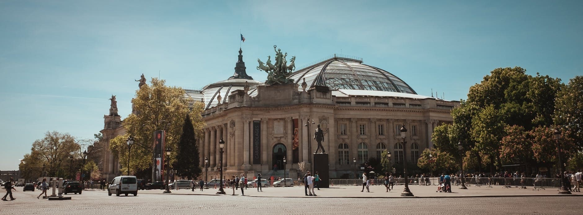 Wide-angle view of the Grand Palais in Paris under a clear blue sky, showcasing its glass dome and ornate architecture with people and cars in the foreground.