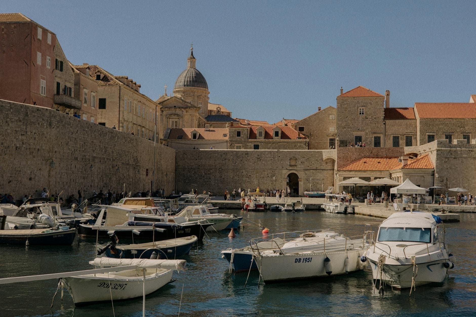 Pequeñas embarcaciones amarradas frente a edificios de piedra estilo Gruž, con el bullicioso mercado y la cúpula de Dubrovnik al fondo bajo un cielo azul despejado.