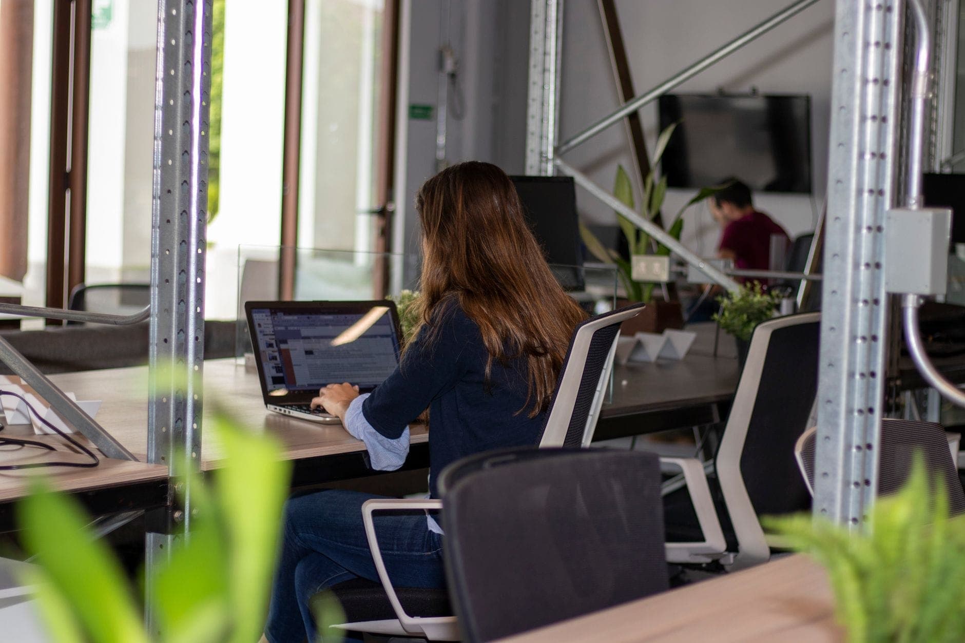 ผู้หญิงทำงานบนแล็ปท็อปในพื้นที่ coworking สมัยใหม่ที่มีต้นไม้และการตกแต่งสไตล์อินดัสเทรียล สื่อถึงบรรยากาศการทำงานที่มีประสิทธิภาพ