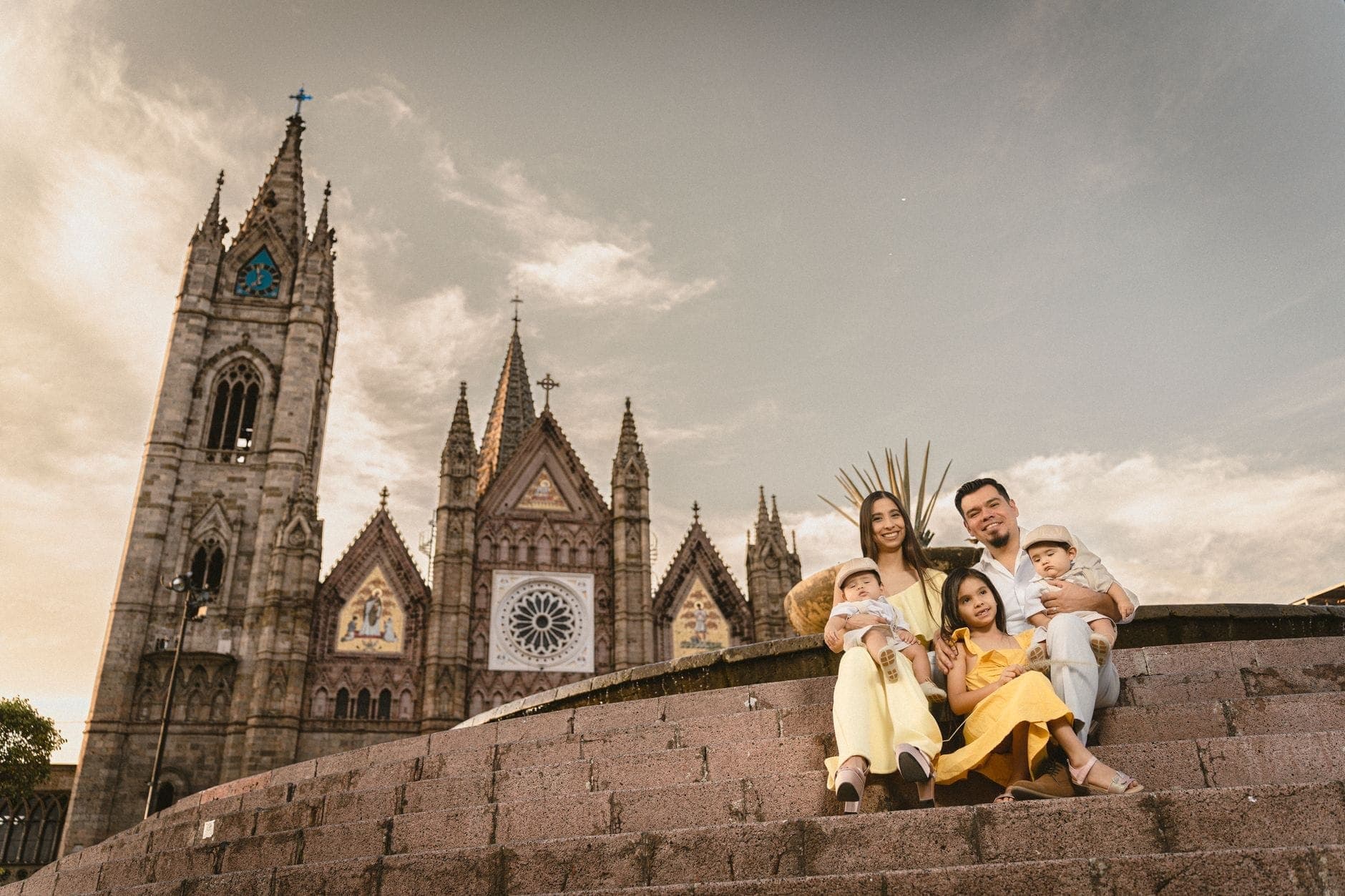 ครอบครัวยิ้มแย้มกับลูกสามคนนั่งบนบันไดหินหน้าวัด Templo Expiatorio ในกวาดาลาฮารา ใต้ท้องฟ้ายามเย็นที่น่าประทับใจ
