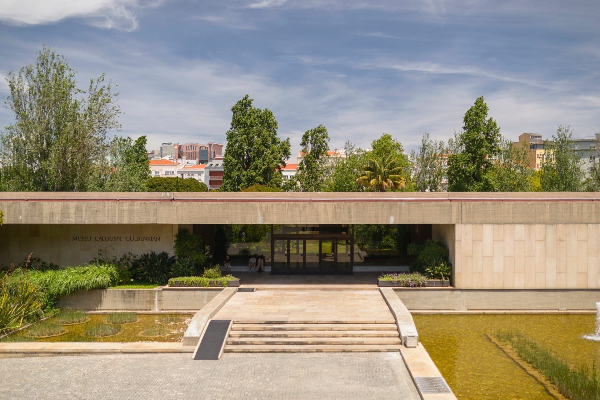 Entrada do Museu Calouste Gulbenkian com fachada moderna em pedra, espelho de água, vegetação exuberante e edifícios da cidade ao fundo sob um céu azul.