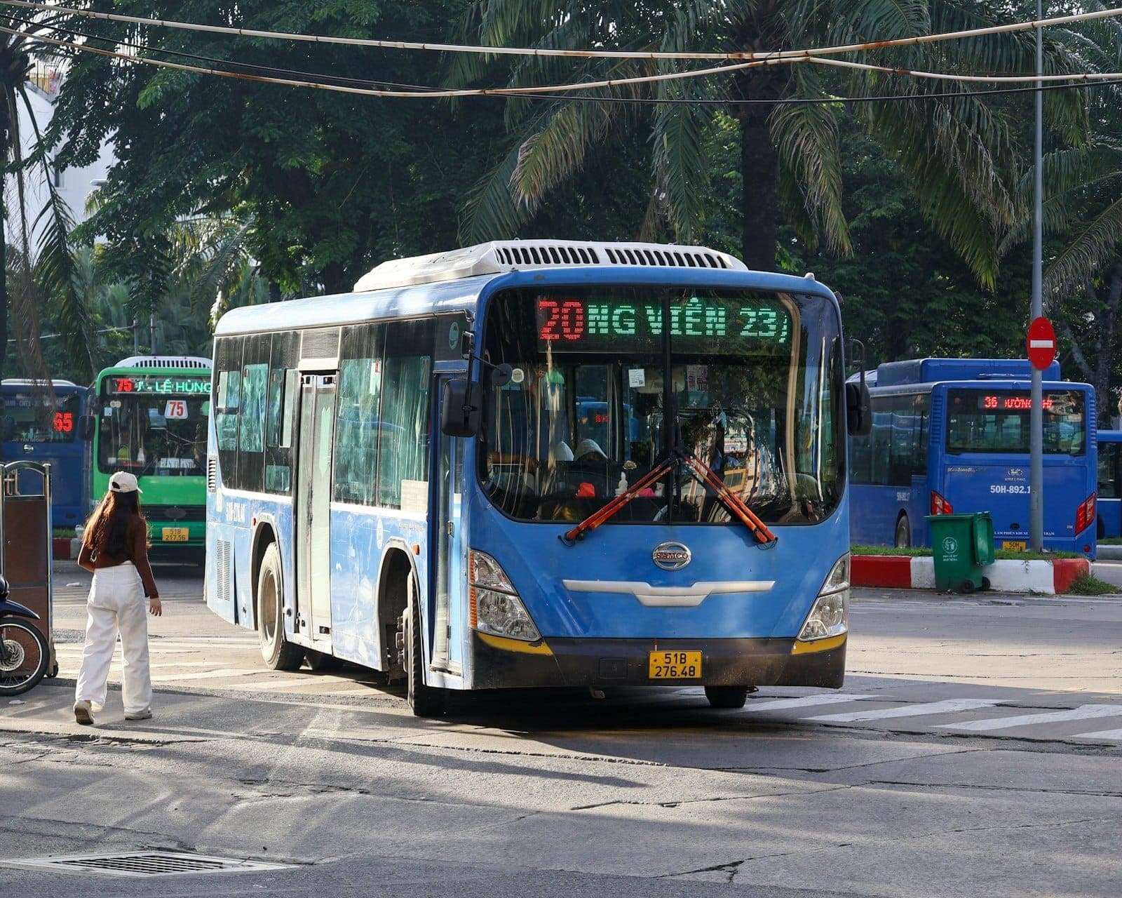 Asul na pampublikong bus ng lungsod sa harap ng mga puno ng palma sa isang interseksyon sa Hanoi, na may aktibidad sa kalye at malinaw na mga karatula.