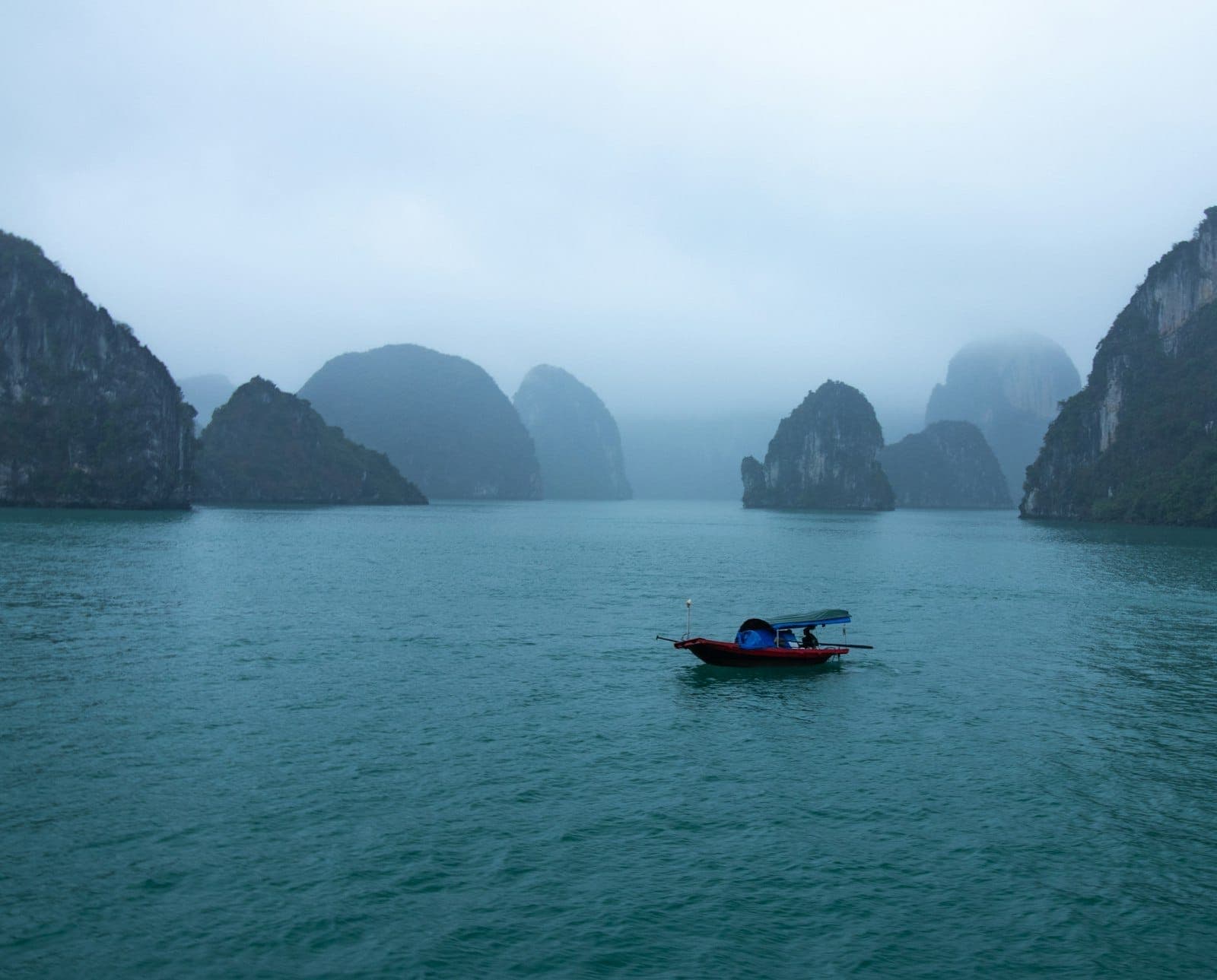 Maliit na bangka sa turquoise na tubig na may mga limestone karst sa likod at maulap na langit sa Ha Long Bay.