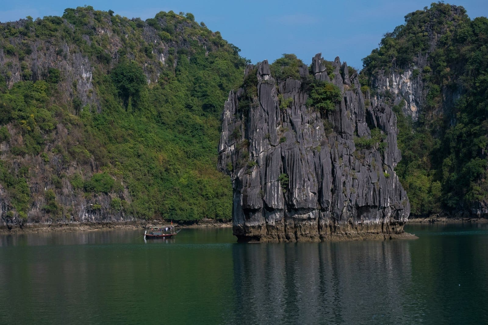 Mataas na limestone karst na batong pormasyon na napapalibutan ng mahinahong berdeng tubig at malusog na mga burol, na may maliit na bangkang kahoy sa tabi.