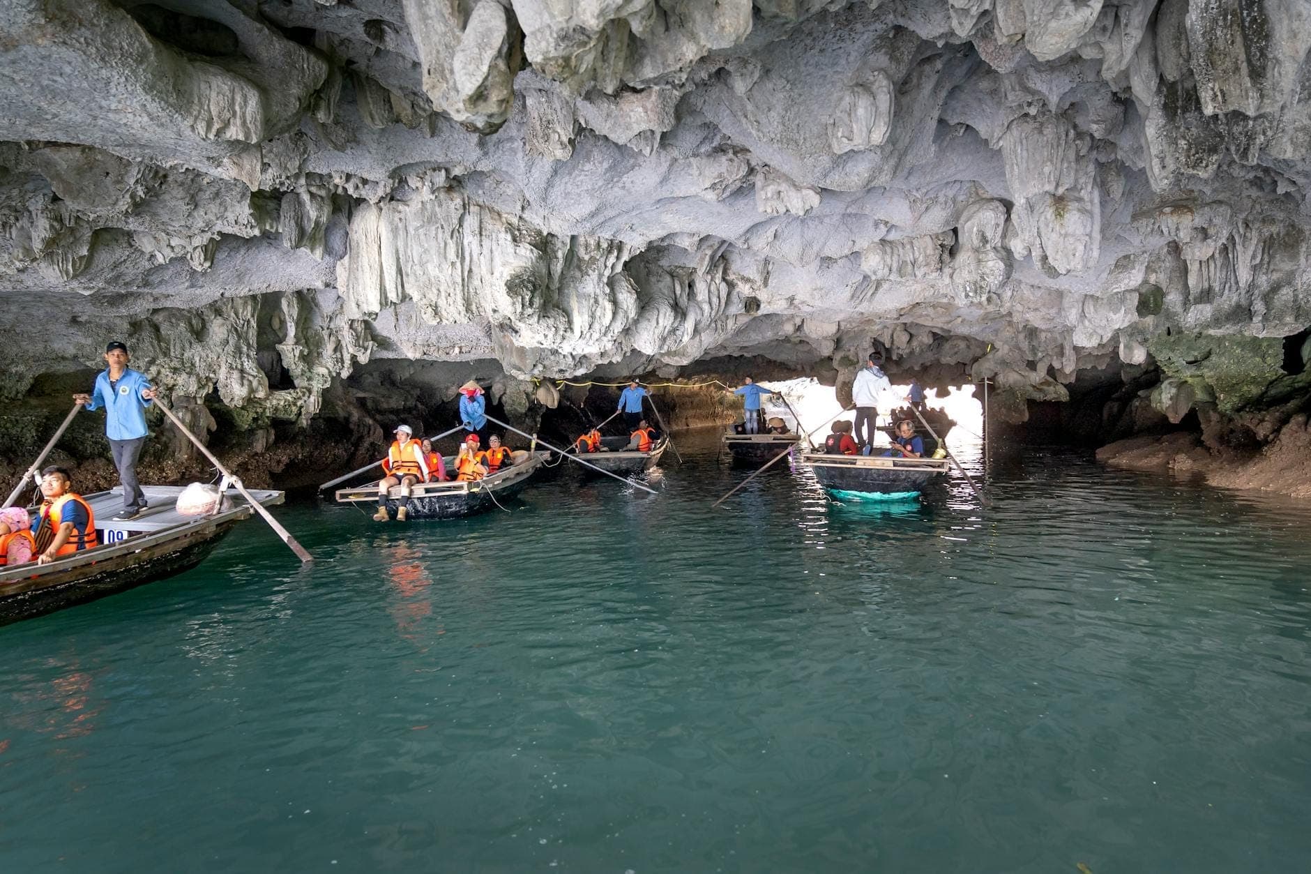 Mga turista sa mga bangkang may sagwan na nakasuot ng life vest na dumadaan sa isang limestone na kuweba sa asul-berdeng tubig sa Ha Long Bay.
