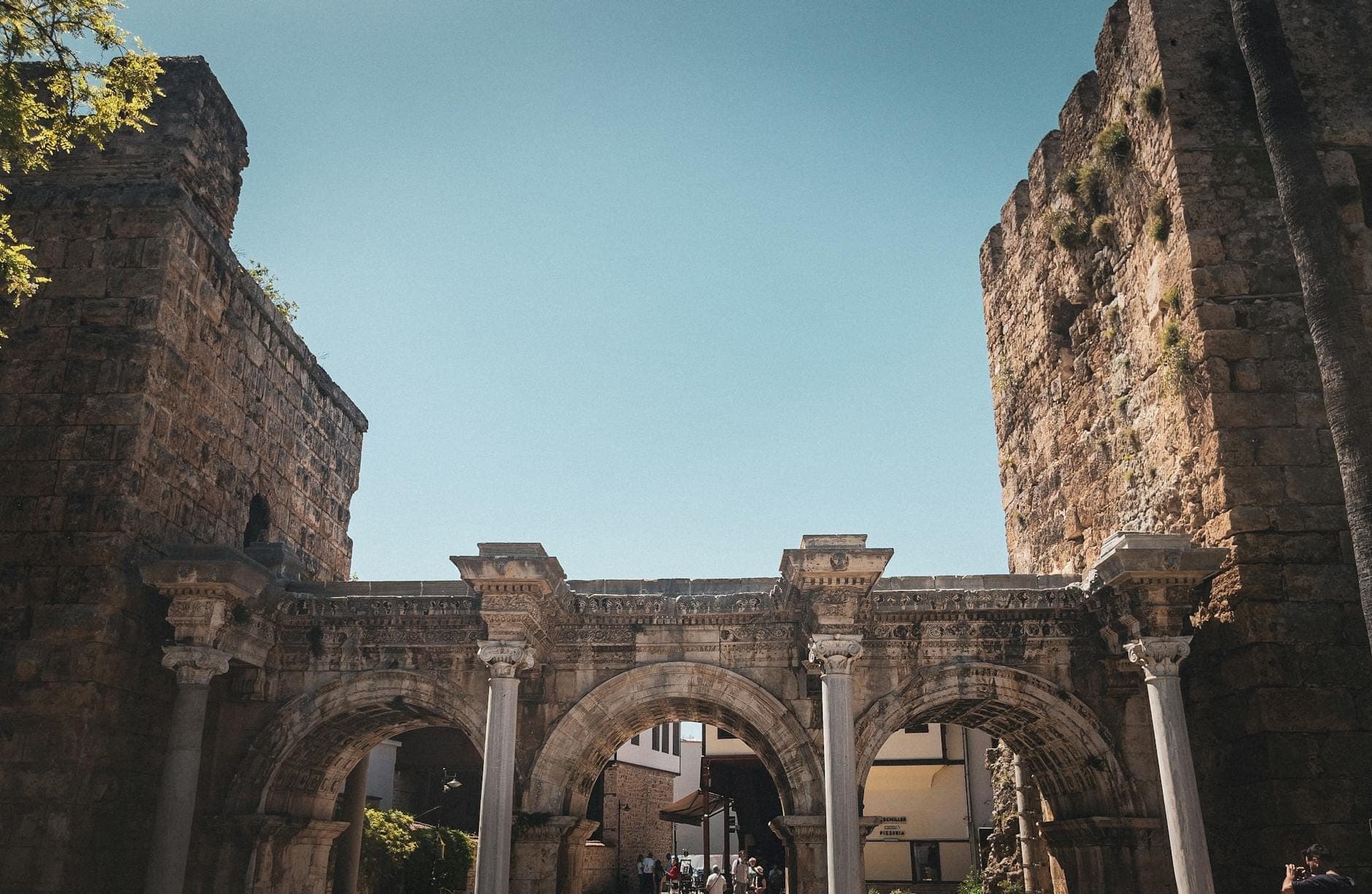 Weitwinkelaufnahme des Hadrianstors in Antalya mit allen drei römischen Marmorbögen, flankiert von antiken Steintürmen unter einem strahlend blauen Himmel.