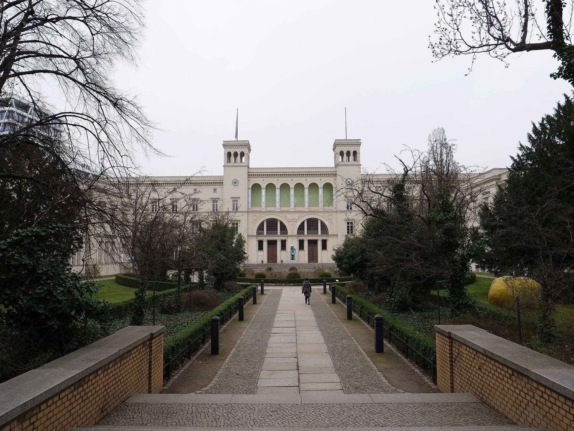 Hamburger Bahnhof в Берлине — величественное неоклассическое здание с двумя башнями, вид с благоустроенной аллеи в серый пасмурный день.