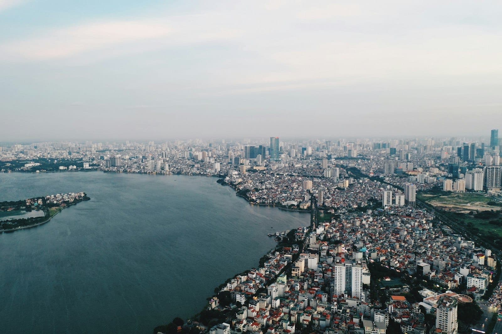 Aerial view of Hanoi cityscape at dusk, showing West Lake and sprawling urban buildings under a soft blue sky, capturing the vastness and essence of Hanoi.