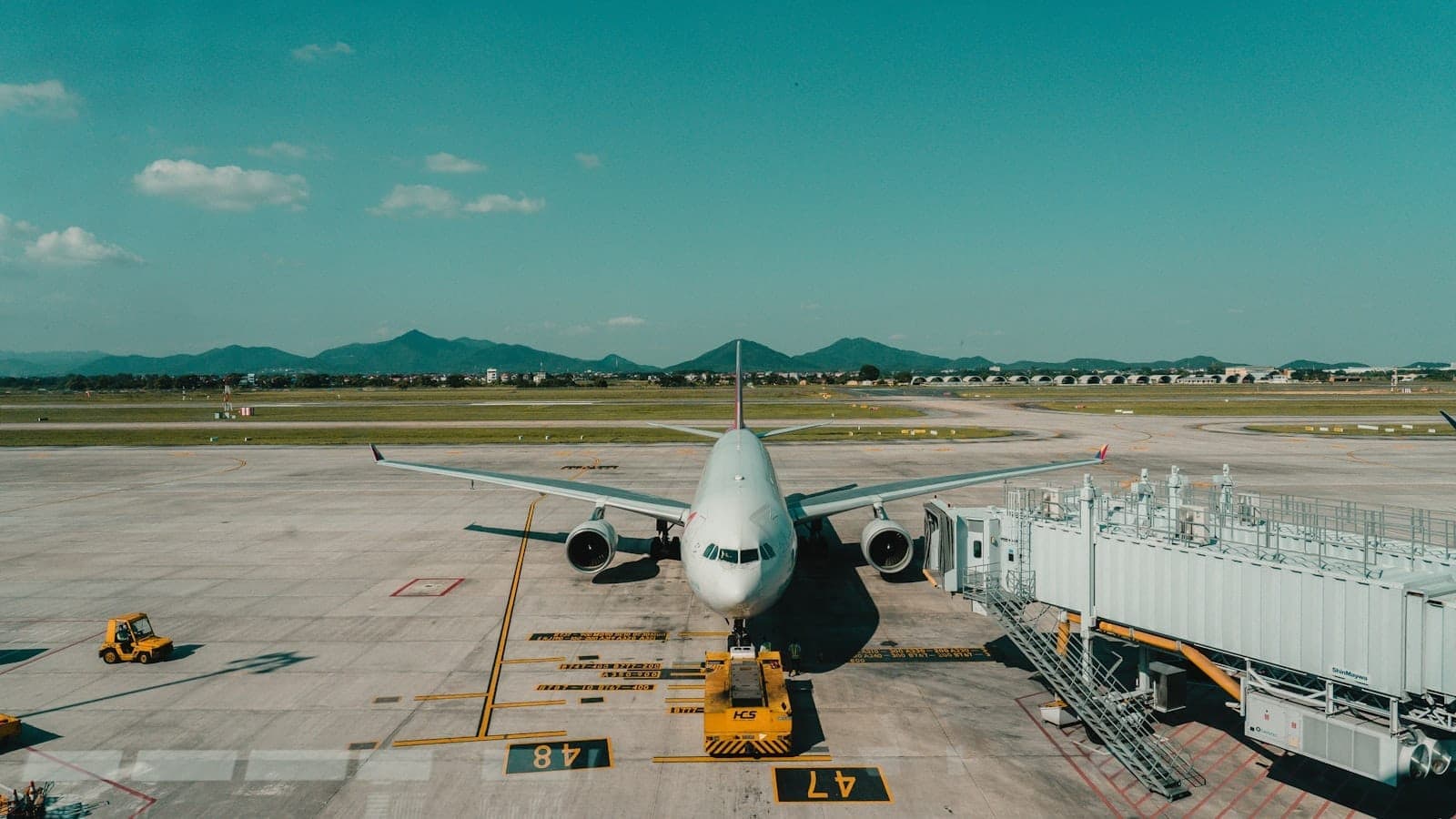 เครื่องบินพาณิชย์ที่เชื่อมต่อกับสะพานเทียบเครื่องบินที่สนามบินนานาชาติ Noi Bai ในฮานอย โดยมีรันเวย์และภูเขาที่อยู่ไกลออกไปให้เห็นภายใต้ท้องฟ้าสีฟ้า