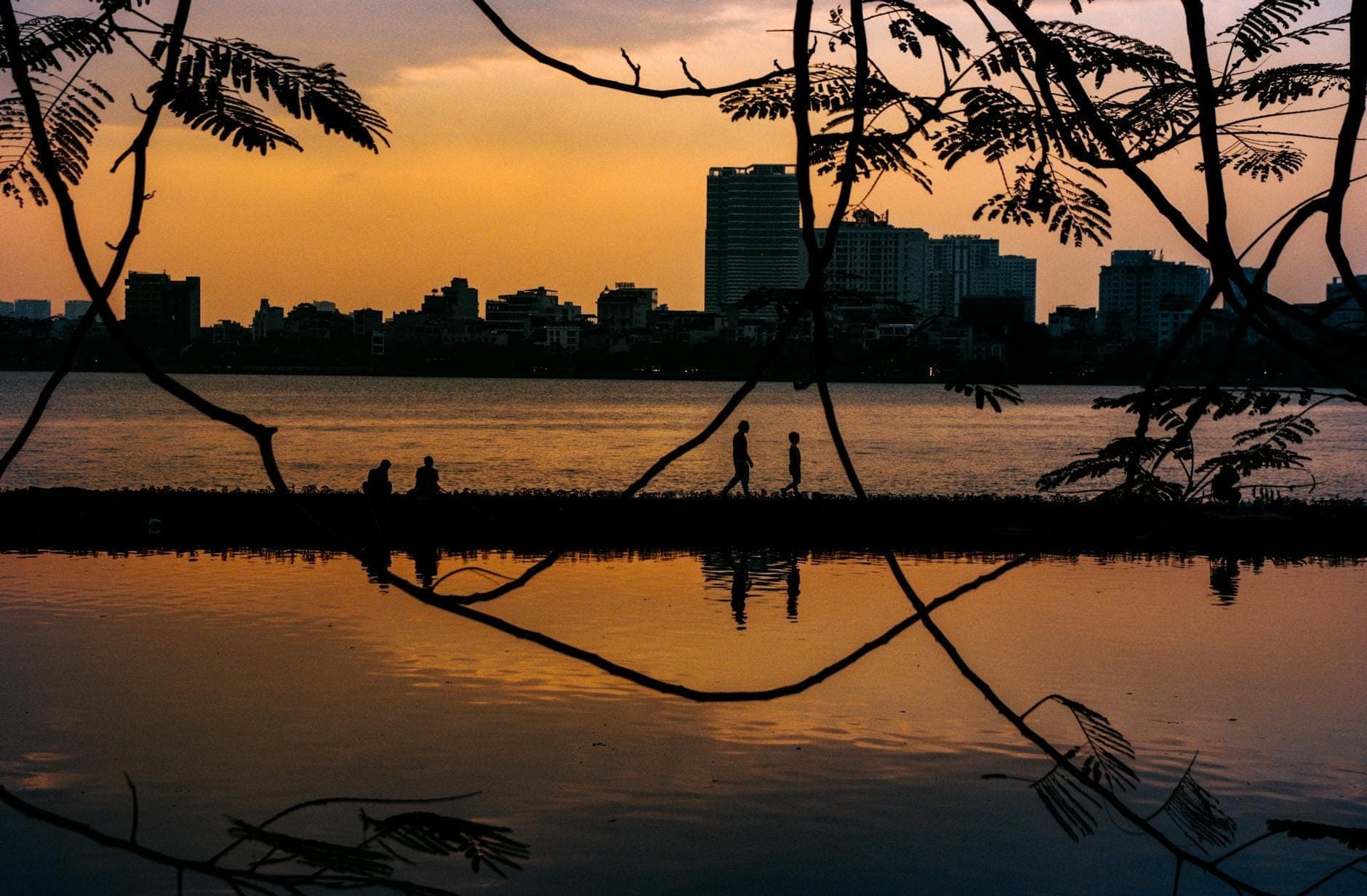เงาของผู้คนที่กำลังเดินและนั่งริมทะเลสาบเตยโห่ในกรุงฮานอยยามพระอาทิตย์ตกดิน โดยมีอาคารในเมืองเป็นฉากหลัง