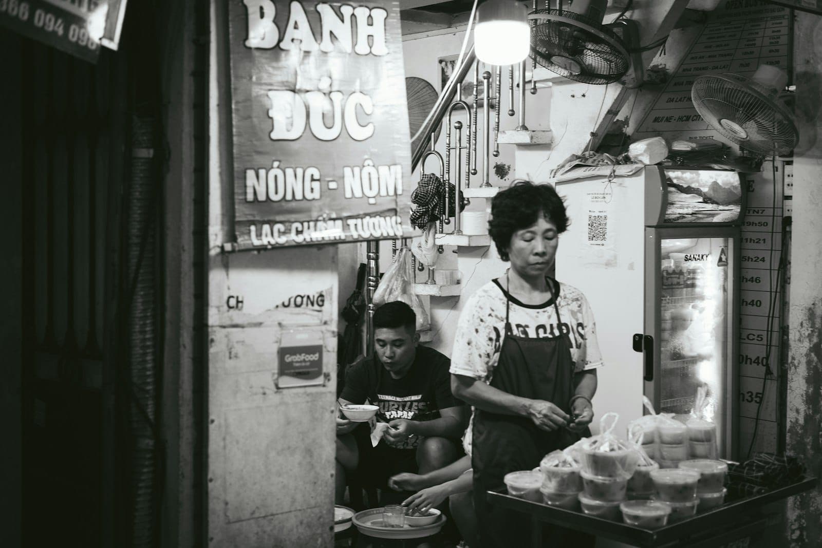 Женщина готовит уличную еду у небольшого лотка в Старом квартале Ханоя, рядом дымящиеся блюда и традиционная вывеска над головой.