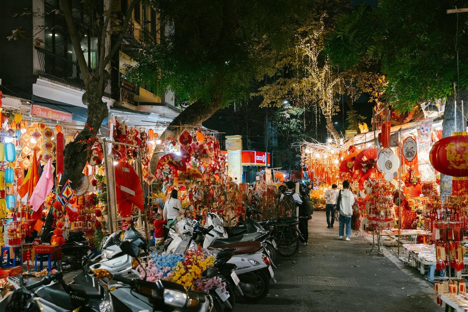 Nocna scena w Starym Kwartale Hanoi z straganami, kolorowymi lampionami, ulicznymi dekoracjami i spacerowiczami wśród zaparkowanych motocykli pod rozświetlonymi drzewami.