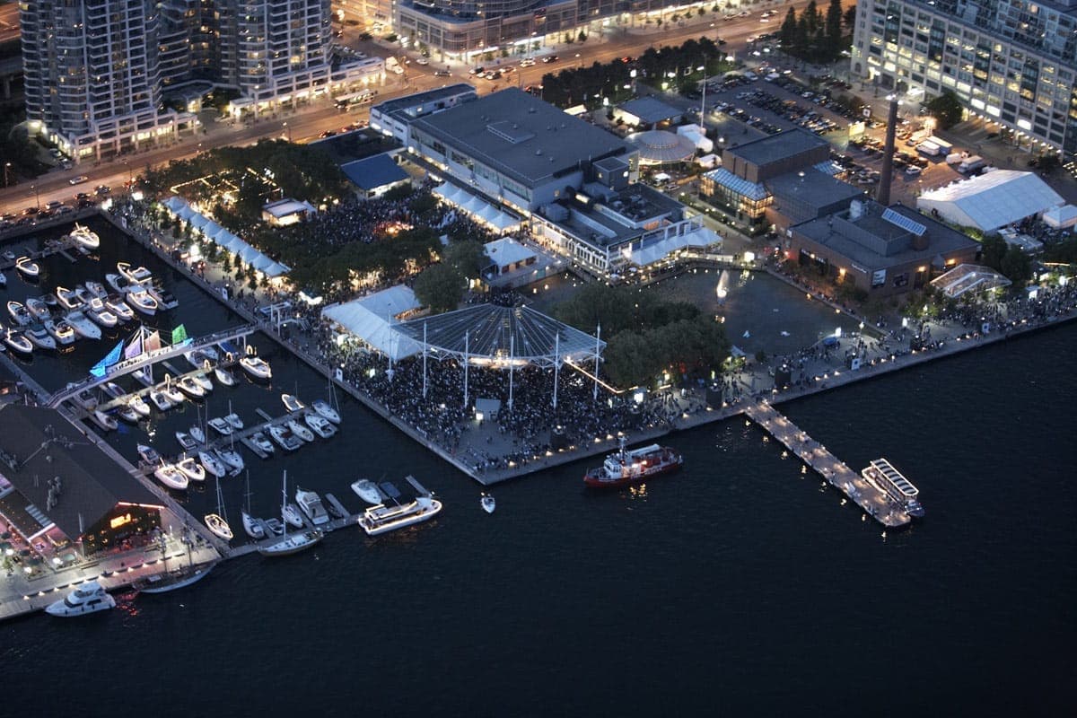 Vista aerea dell'Harbourfront Centre a Toronto al crepuscolo, con edifici sul lungomare, tendoni per eventi, un porto turistico con barche e folle che godono della serata.