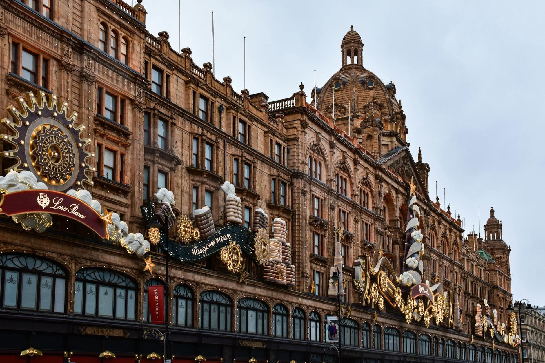 Vista panorámica de la icónica fachada histórica de Harrods en Londres, adornada con decoraciones festivas y detalles dorados bajo un cielo nublado, que captura la grandiosidad del almacén y su ambiente navideño.