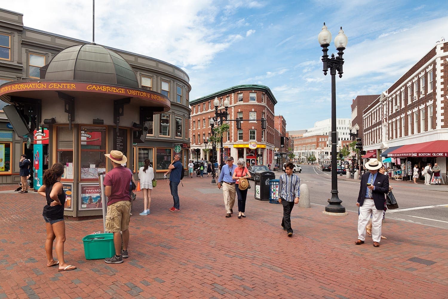 Des personnes se promènent et se rassemblent près du Cambridge Visitor's Information Center à Harvard Square, entourées de bâtiments historiques en briques sous un ciel partiellement nuageux.