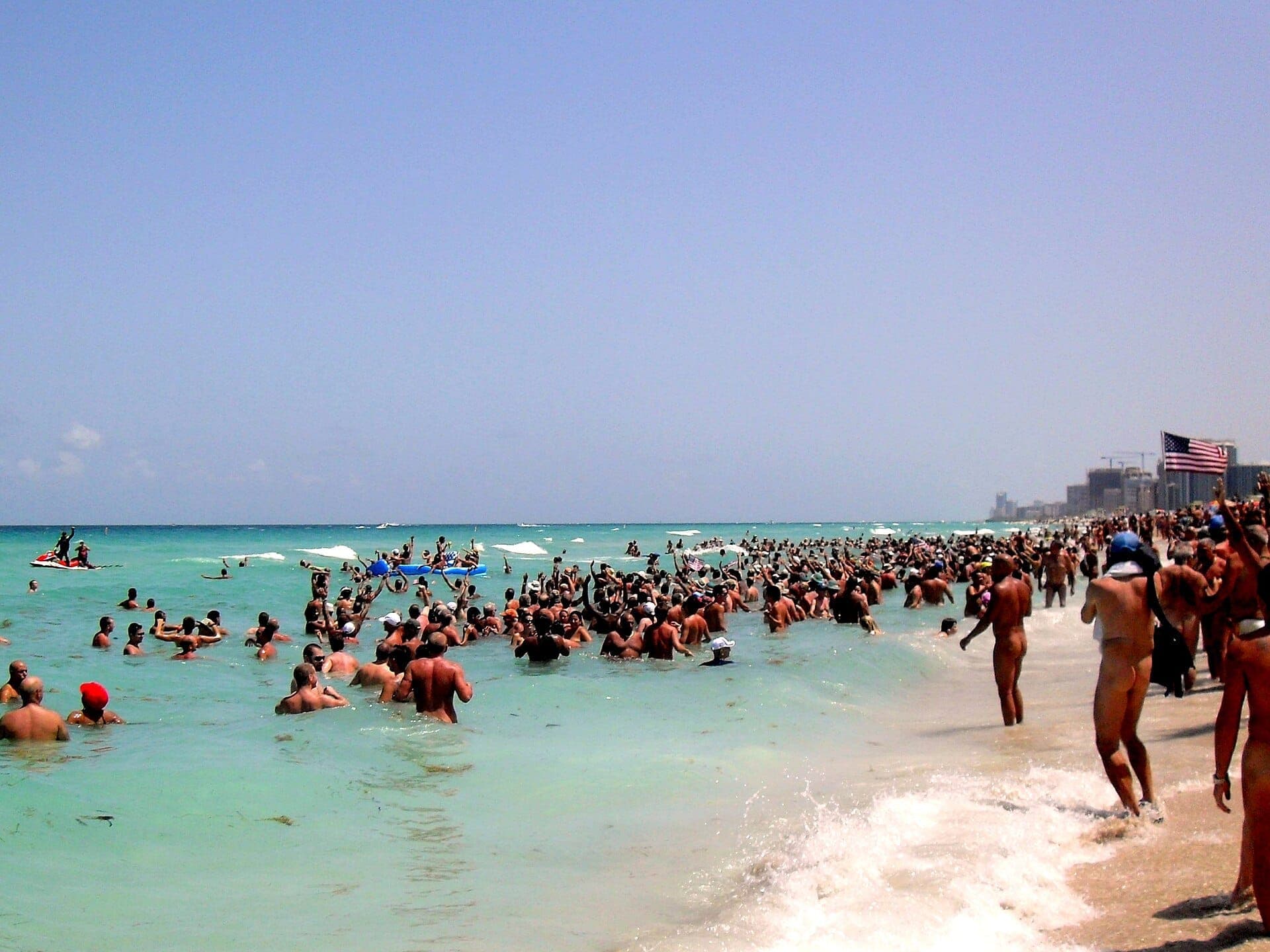 Crowds enjoy the clear turquoise water and sandy shore at Haulover Beach Park under a bright blue sky, with city buildings in the distance.