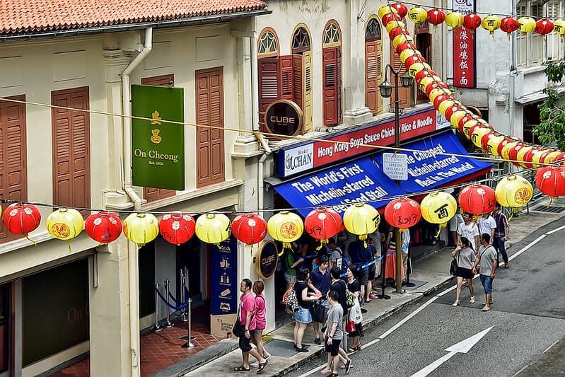 Vista noturna de pessoas na fila em uma barraca de comida bem iluminada, com carnes assadas penduradas, em um hawker center de Singapura.