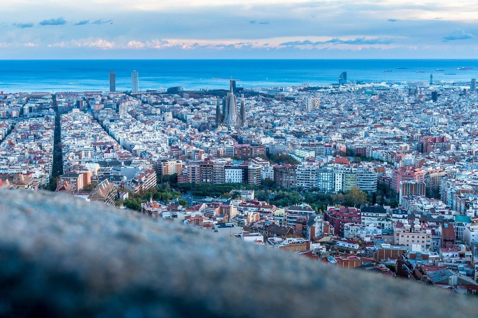 ภาพถ่ายทางอากาศมุมกว้างของบาร์เซโลนายามพลบค่ำ โดดเด่นด้วย Sagrada Família อันเป็นสัญลักษณ์ ถนนแบบตาราง อาคารสีสันสดใส และทะเลเมดิเตอร์เรเนียนในระยะไกล