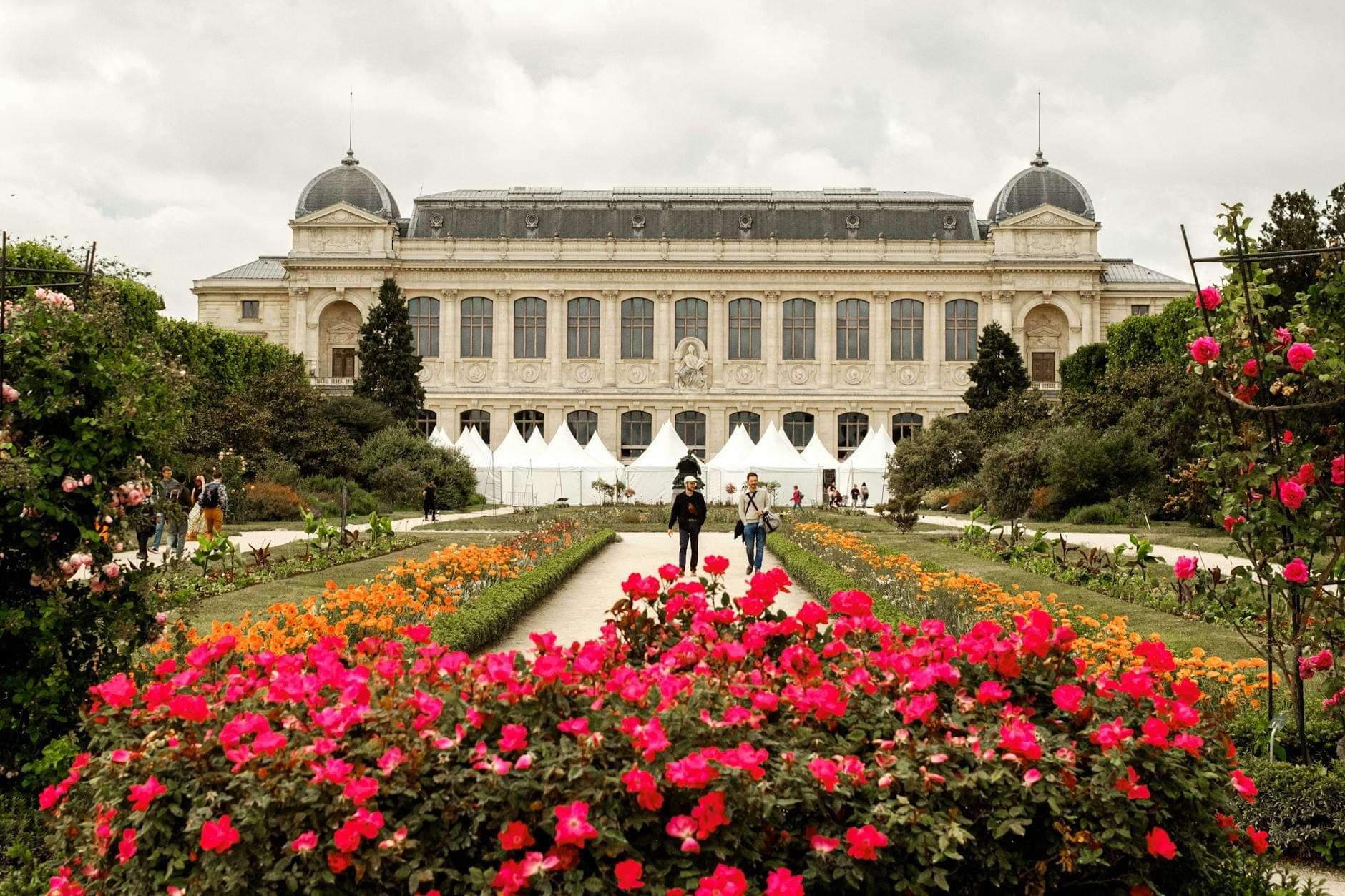 Elegante Fassade eines historischen Museums in Paris hinter üppigen Gärten mit bunten Blumen und Wegen, Menschen schlendern draußen.