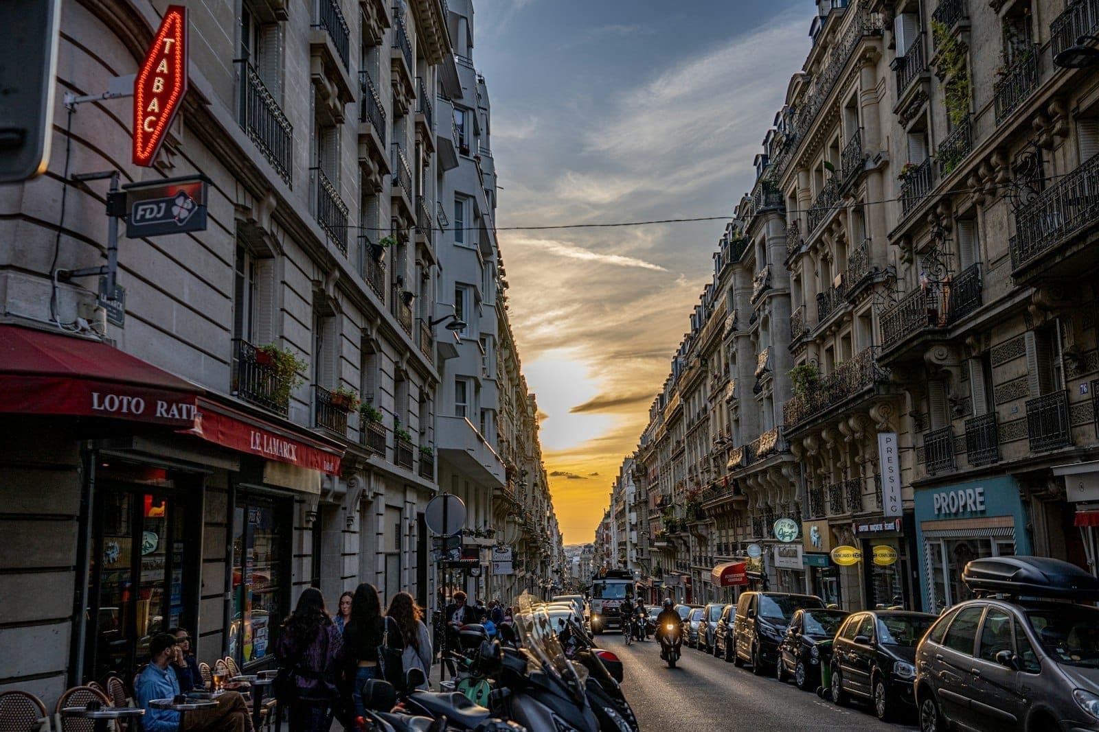 A lively Parisian street at sunset with people, scooters, and classic buildings lining both sides under a dramatic evening sky.