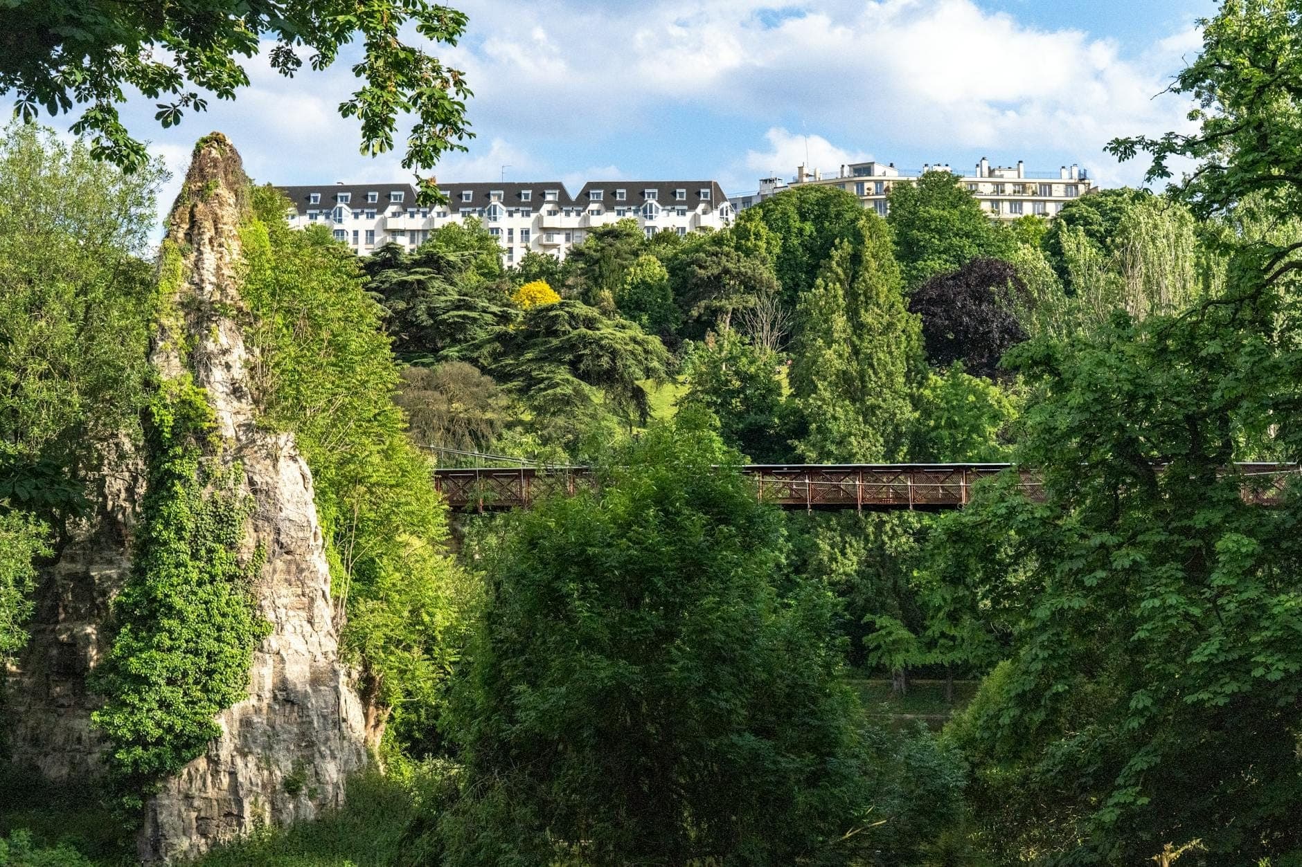 Üppiger grüner Park mit dramatischem Felsvorsprung, Fußgängerbrücke und Pariser Gebäuden im Hintergrund unter blauem Himmel.