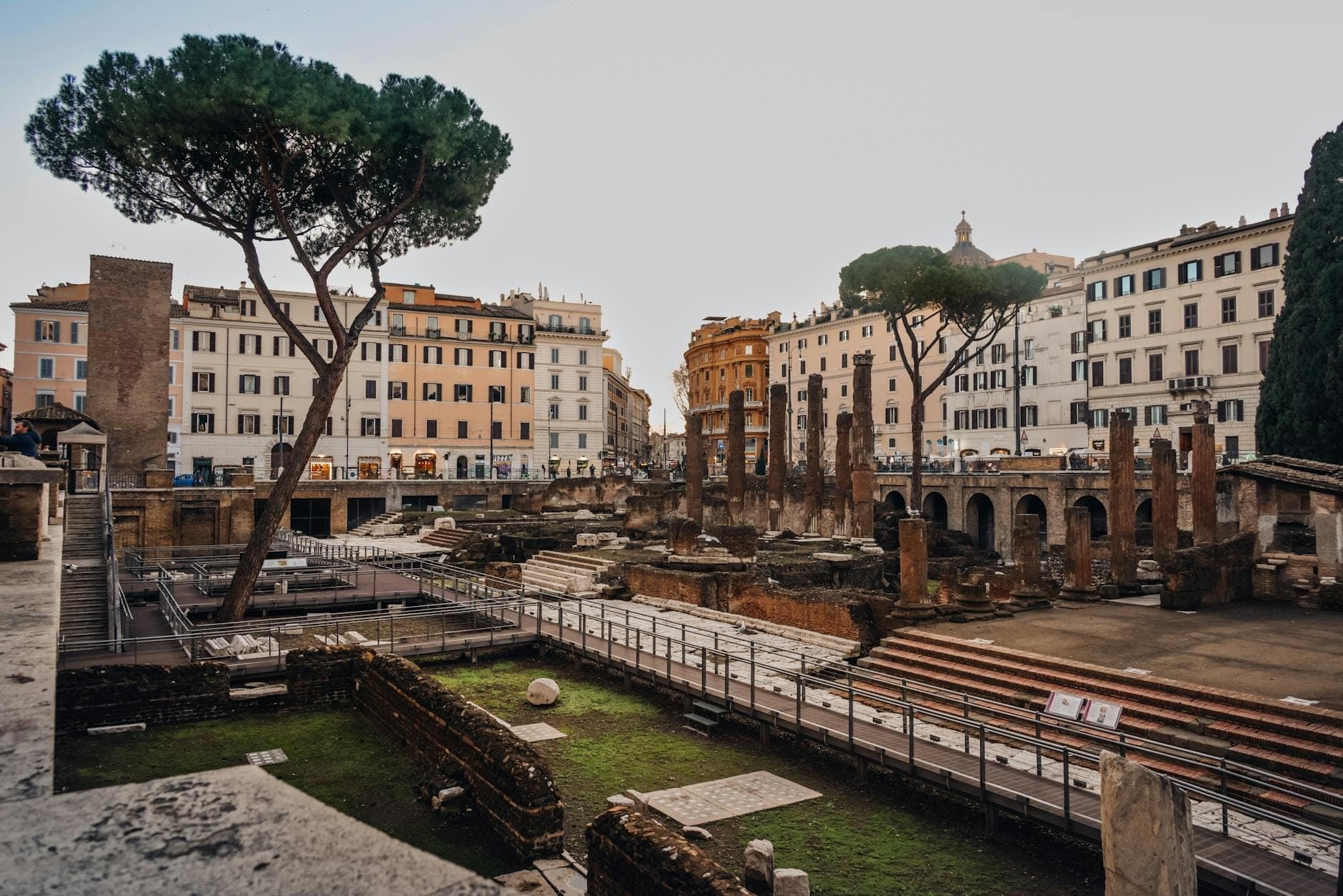 Pemandangan reruntuhan Romawi kuno dengan kolom-kolom dan penggalian bawah tanah, dikelilingi bangunan khas Roma di pusat kota Roma.