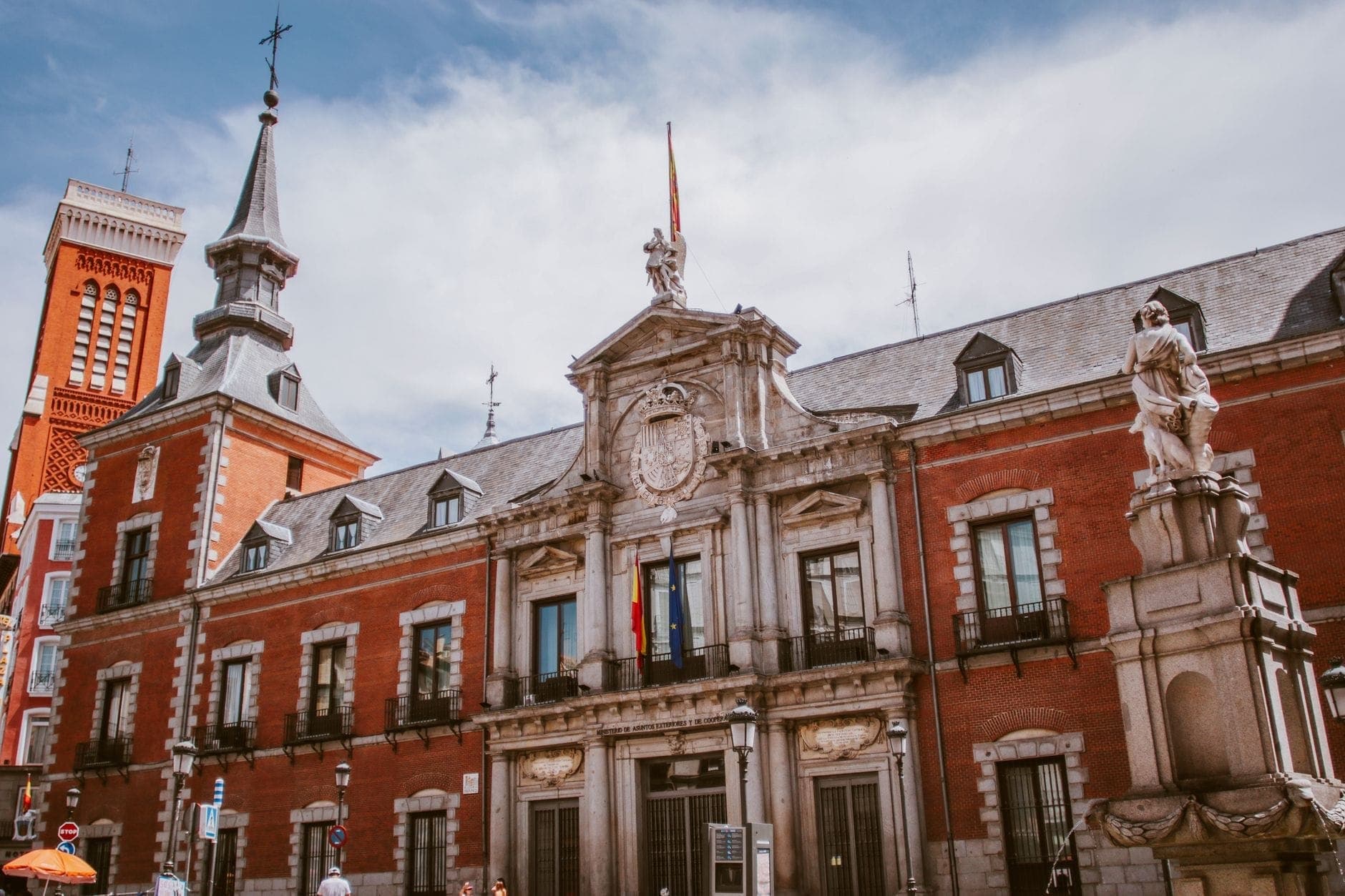 Edifício histórico de tijolo e pedra em Madrid com detalhes arquitetônicos ornamentados e uma estátua na frente, sob um céu parcialmente nublado.