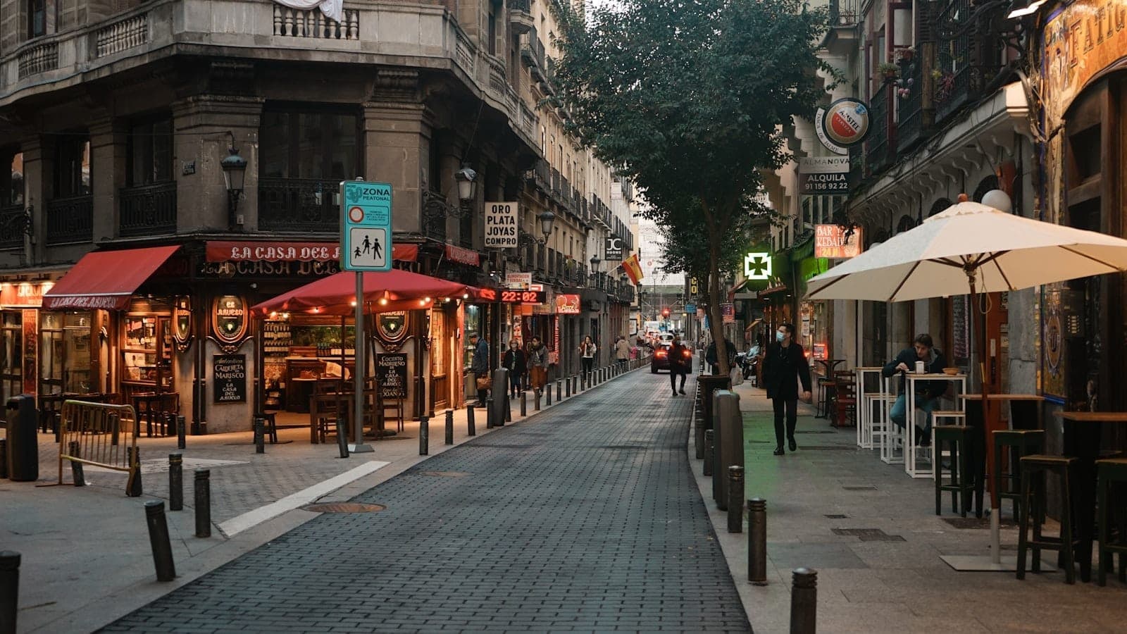 Uma movimentada rua de Madrid repleta de cafés e restaurantes, mesas ao ar livre, pedestres e arquitetura clássica, capturando perfeitamente a cena gastronômica de um bairro local.