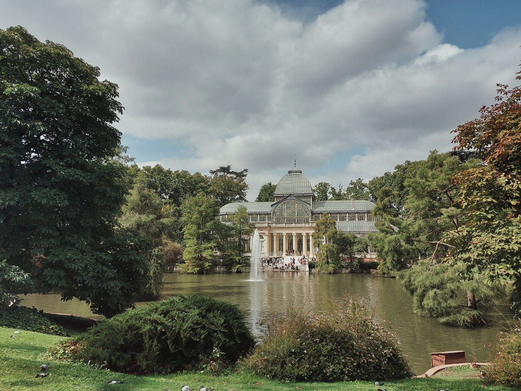Uma cena tranquila de jardim com o envidraçado Palacio de Cristal, um lago, vegetação ao redor e uma fonte no Parque del Retiro de Madrid.