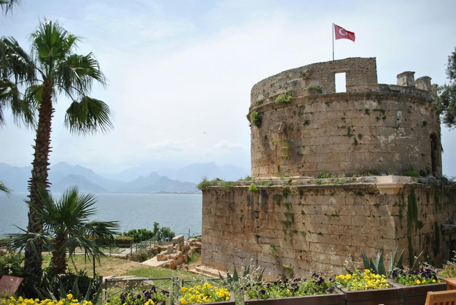 Der Hıdırlık-Turm, ein runder römischer Steinturm mit einer türkischen Flagge auf der Spitze, erhebt sich über dem Meer, im Hintergrund Berge und Palmen.