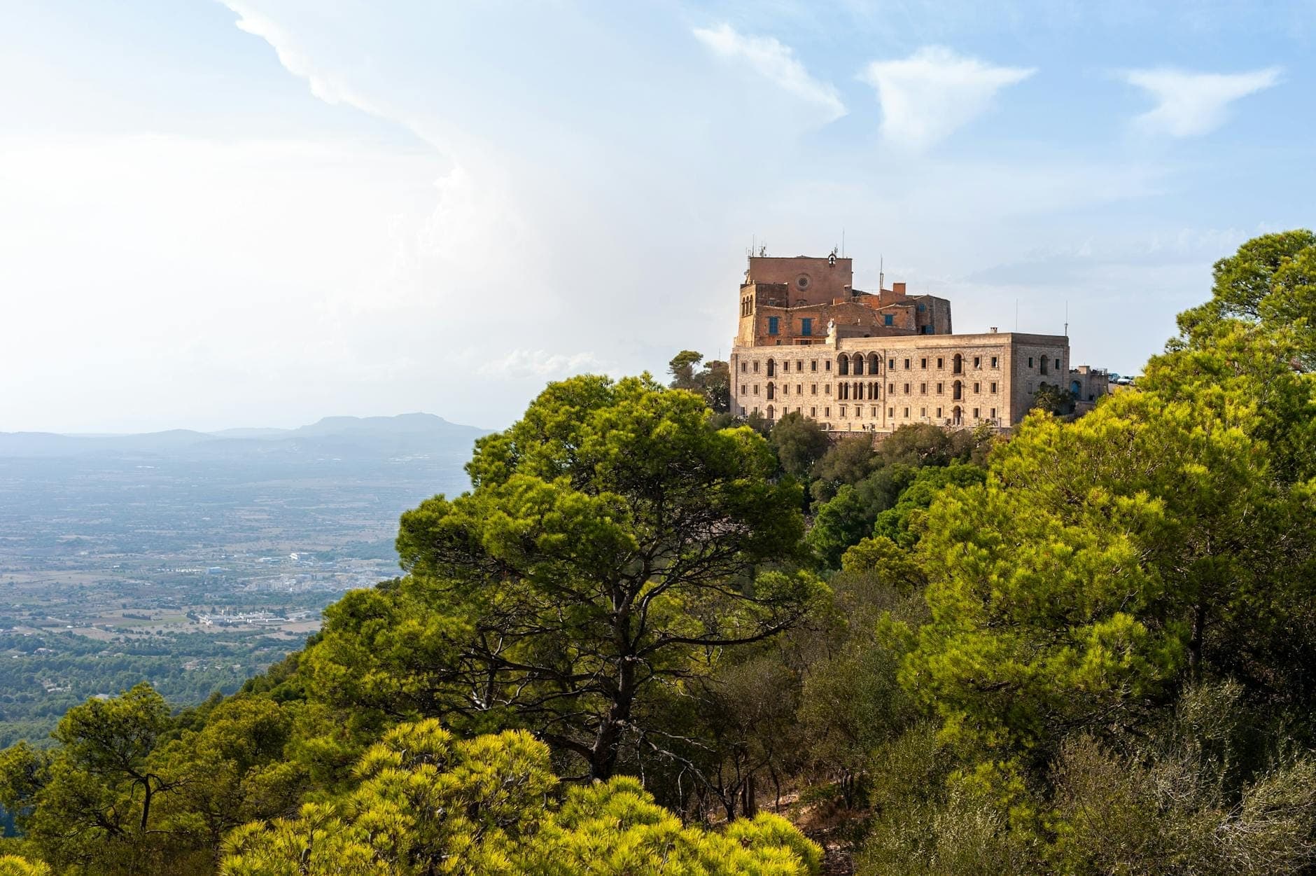 Storico edificio monastico su una collina circondata da pini che si affaccia sulle pianure di Mallorca, con un cielo sereno sopra.