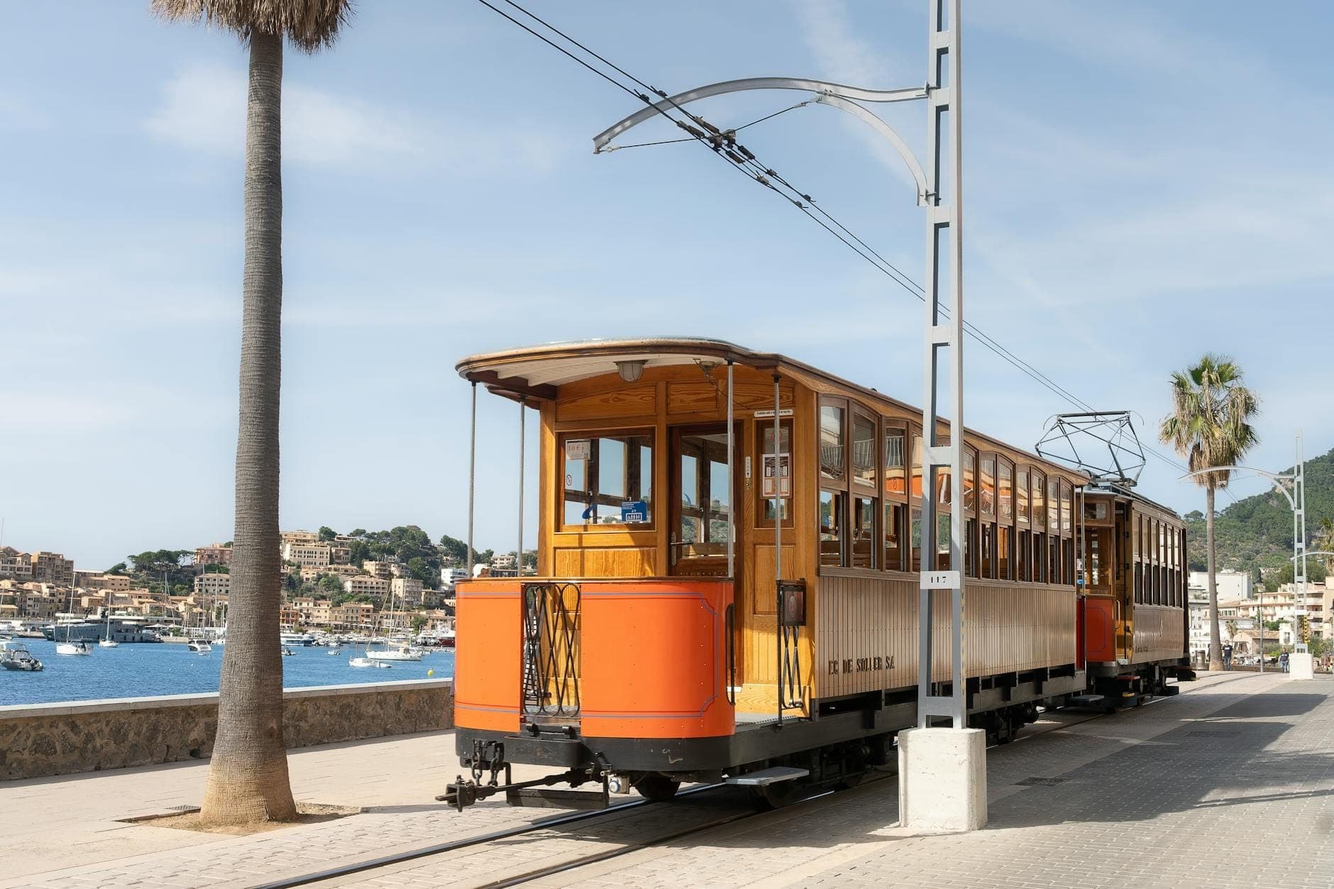 Storico tram in legno sul lungomare di Port de Sóller, Mallorca, con montagne e villaggio sullo sfondo.