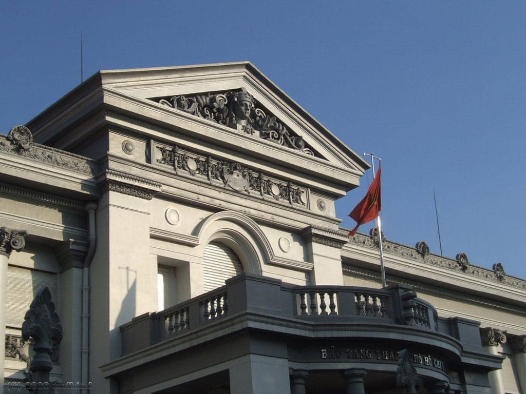 Harapang fasad ng Museum ng Ho Chi Minh na may arkitekturang kolonyal ng Pransya, masalimuot na ukit sa bato, at bandilang Vietnamese sa ilalim ng maliwanag na asul na langit.