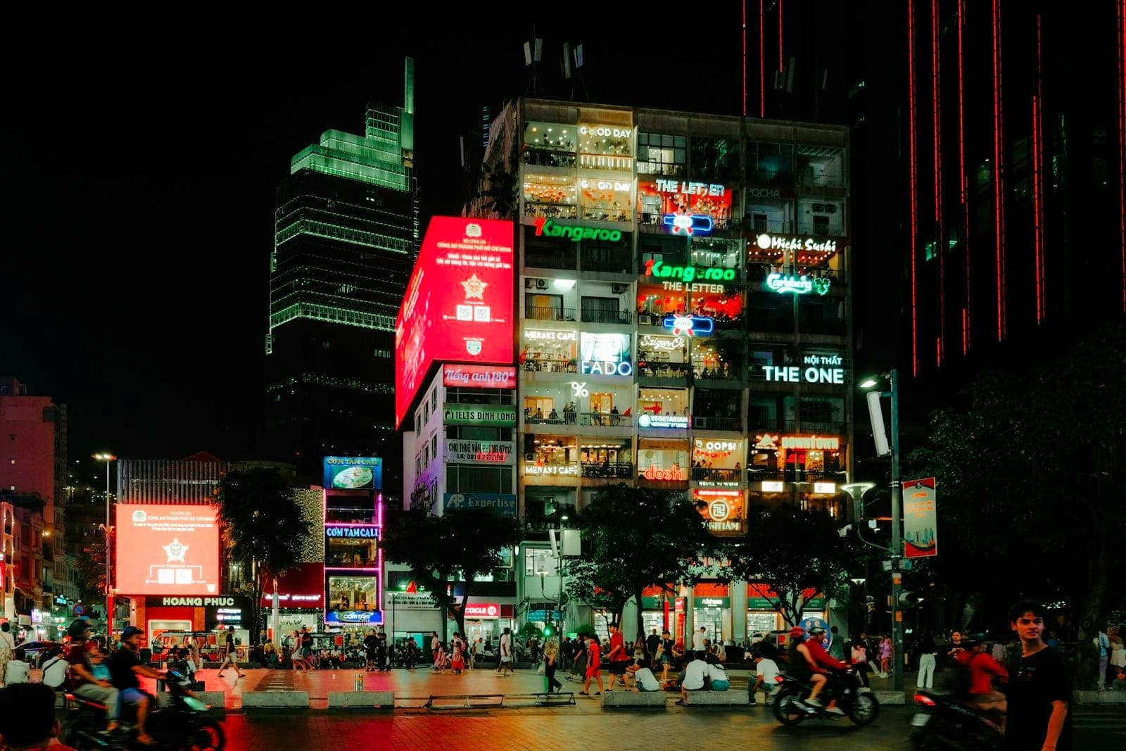 Ho Chi Minh City'de pencerelerden görünen birden fazla aydınlatılmış bar ve lounge bulunan bir bina, önde gece insanlar ve scooterlarla.