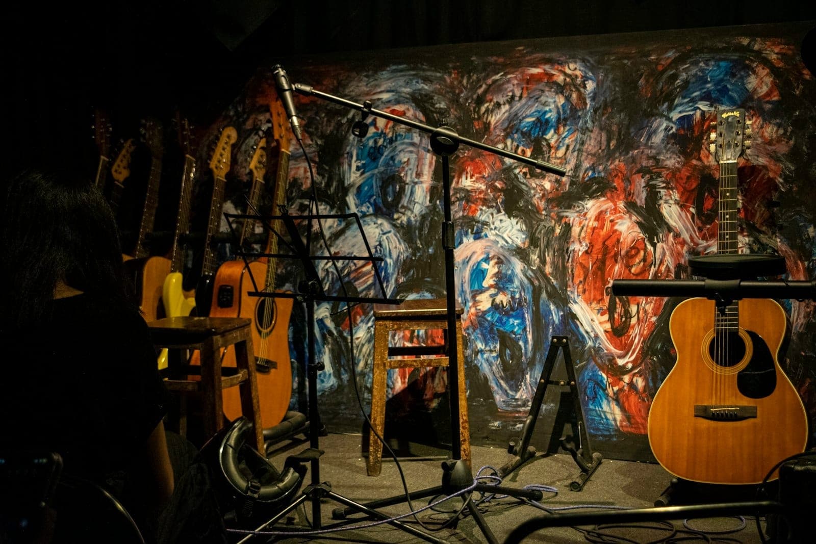 Birden fazla gitar, mikrofon standı, tabure ve renkli soyut duvar resmiyle süslenmiş arka plana sahip, performansa hazır samimi bir canlı müzik sahnesi.