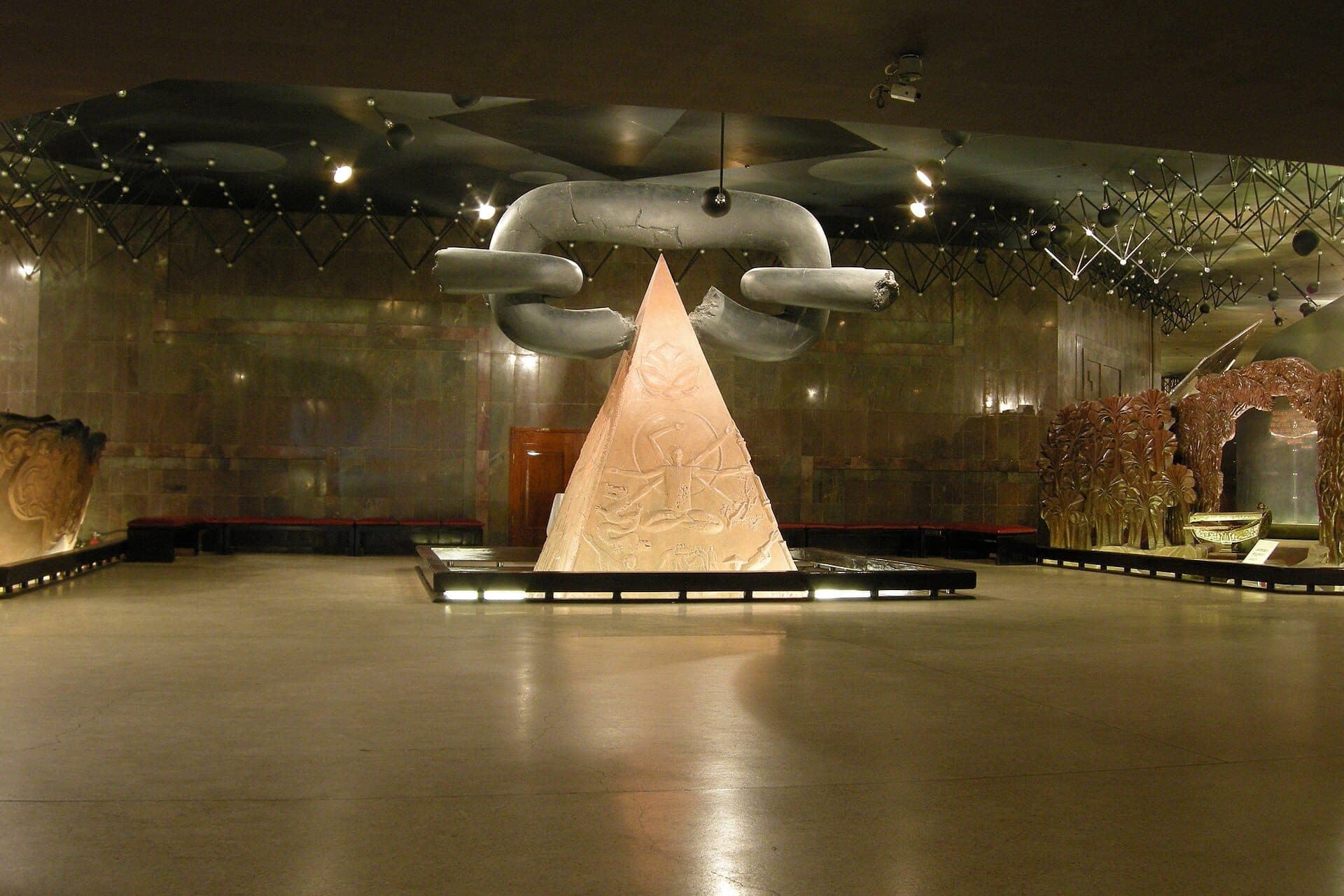 Główna sala Muzeum Ho Chi Minh z dużą rzeźbą w kształcie piramidy i ogromnymi metalowymi ogniwami łańcucha w dramatycznym oświetleniu.