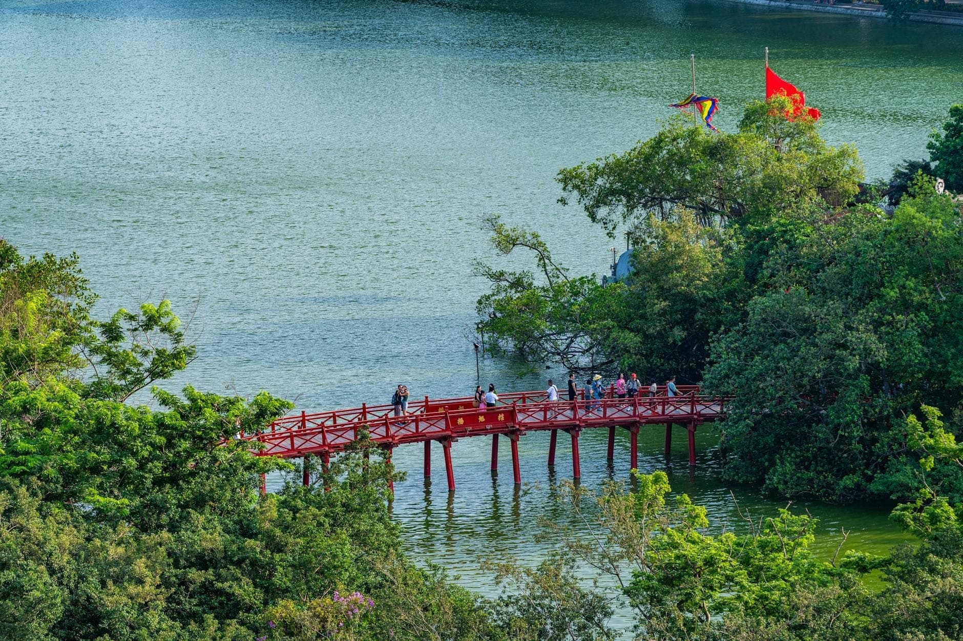 Widok z lotu ptaka na jezioro Hoàn Kiếm z charakterystycznym czerwonym mostem Thê Húc prowadzącym nad wodą ku porośniętej drzewami świątyni, otoczonej bujną zielenią i wietnamskimi flagami.