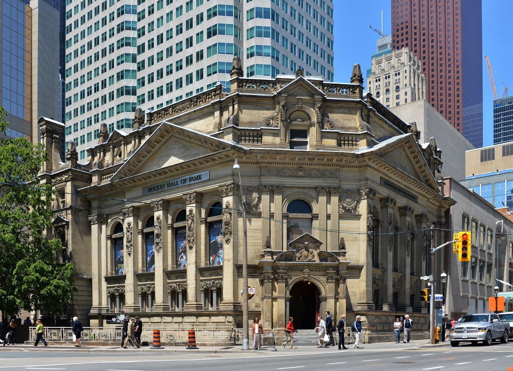 Vista esterna dell'Hockey Hall of Fame, uno storico edificio bancario dell'Ottocento nel centro di Toronto, con persone che camminano fuori e grattacieli moderni sullo sfondo.