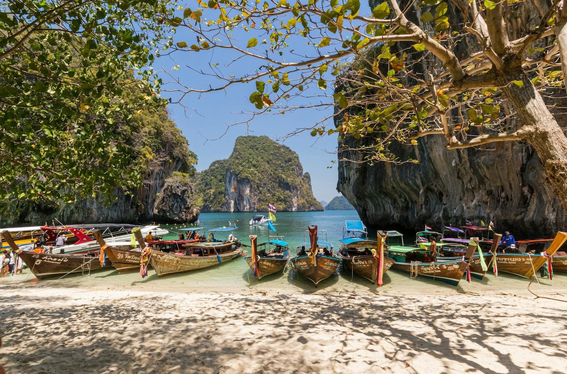 Scenic view from shaded sandy beach with traditional longtail boats moored in turquoise water, dramatic limestone cliffs, and lush islands in the background under a clear sky.
