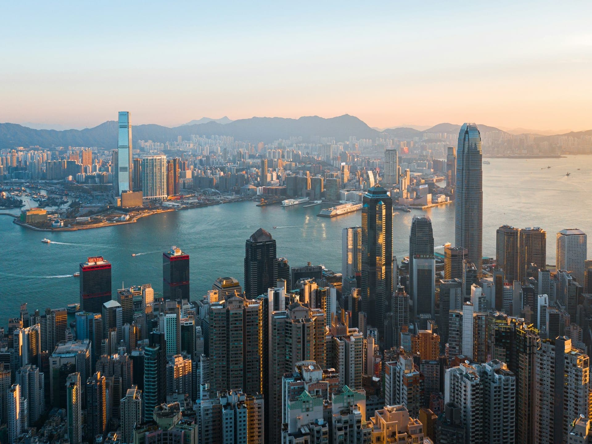 Hong Kong skyline at sunset overlooking Victoria Harbour with skyscrapers, waterfront, and mountains in the background