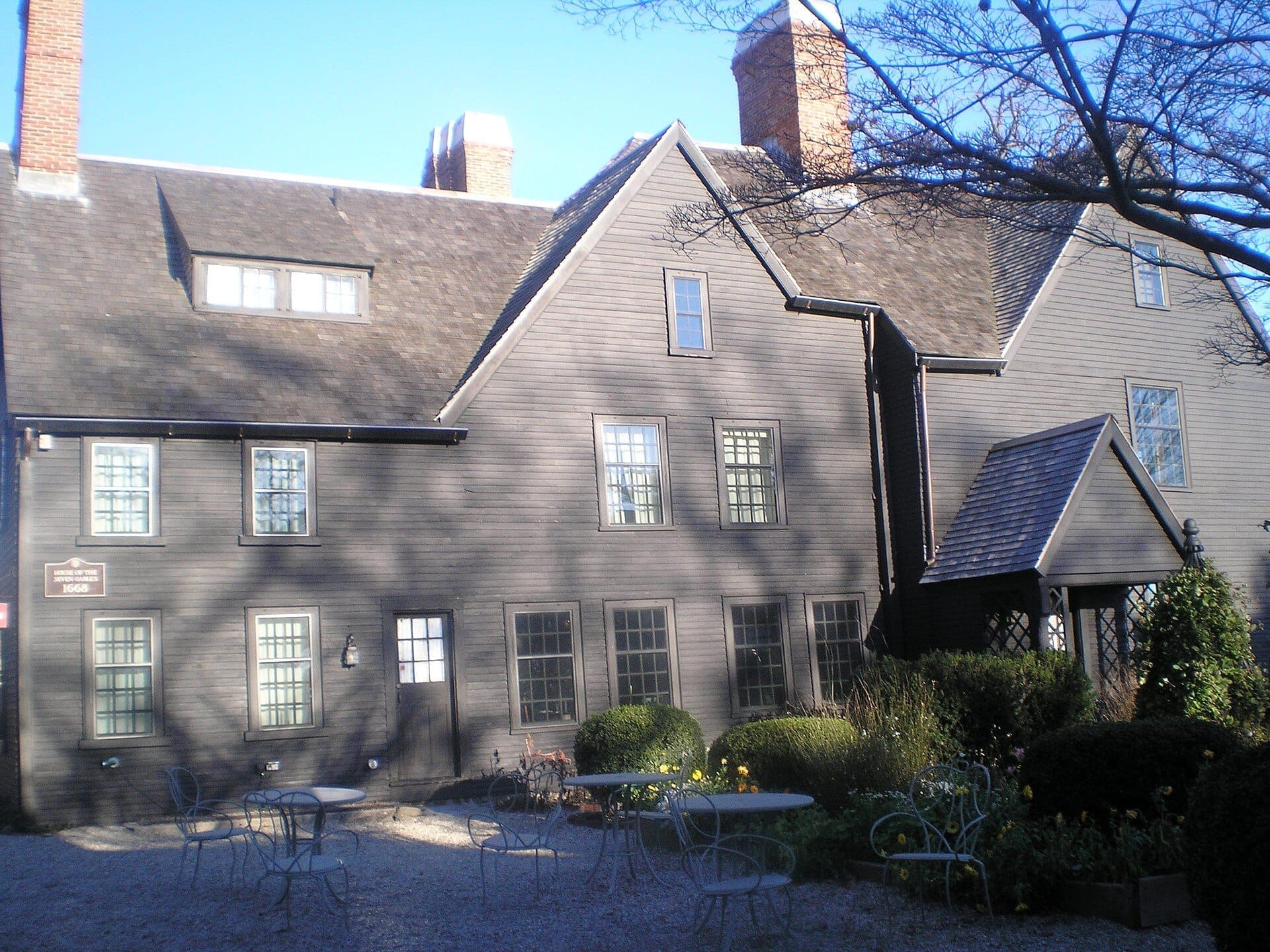 La Maison aux Sept Pignons à Salem, un manoir historique en bois sombre avec plusieurs pignons, des cheminées en brique et un espace salon dans le jardin extérieur.