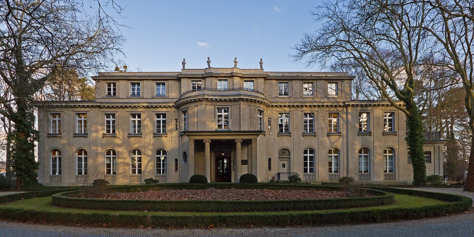 มุมมองด้านหน้าของ House of the Wannsee Conference วิลล่าประวัติศาสตร์สีเบจพร้อมหน้าต่างที่สมมาตรและสวนที่ตกแต่งอย่างดีในวันที่อากาศแจ่มใส