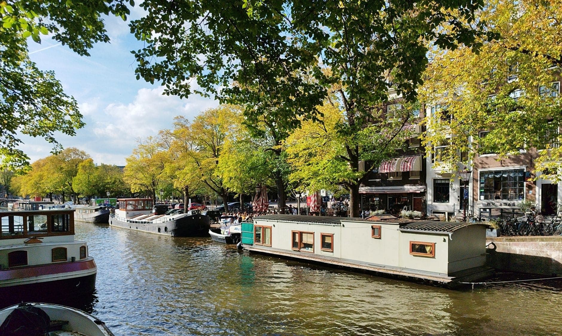 Eine Reihe bunter Hausboote liegt an einer Amsterdamer Gracht, gesäumt von üppigen Bäumen und historischen Backsteingebäuden an einem sonnigen Tag.
