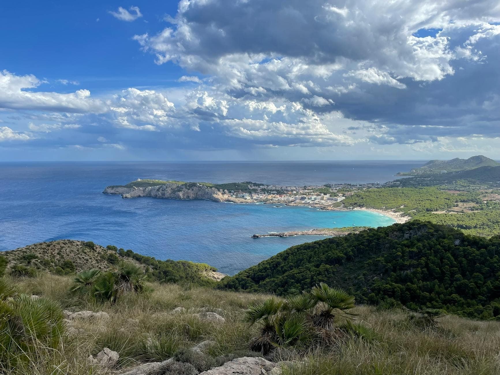 Weiter Blick auf die Küste Mallorcas mit grünen Hügeln, einem Strand und wechselnden Wolken, die unterschiedliche Wettermuster andeuten.