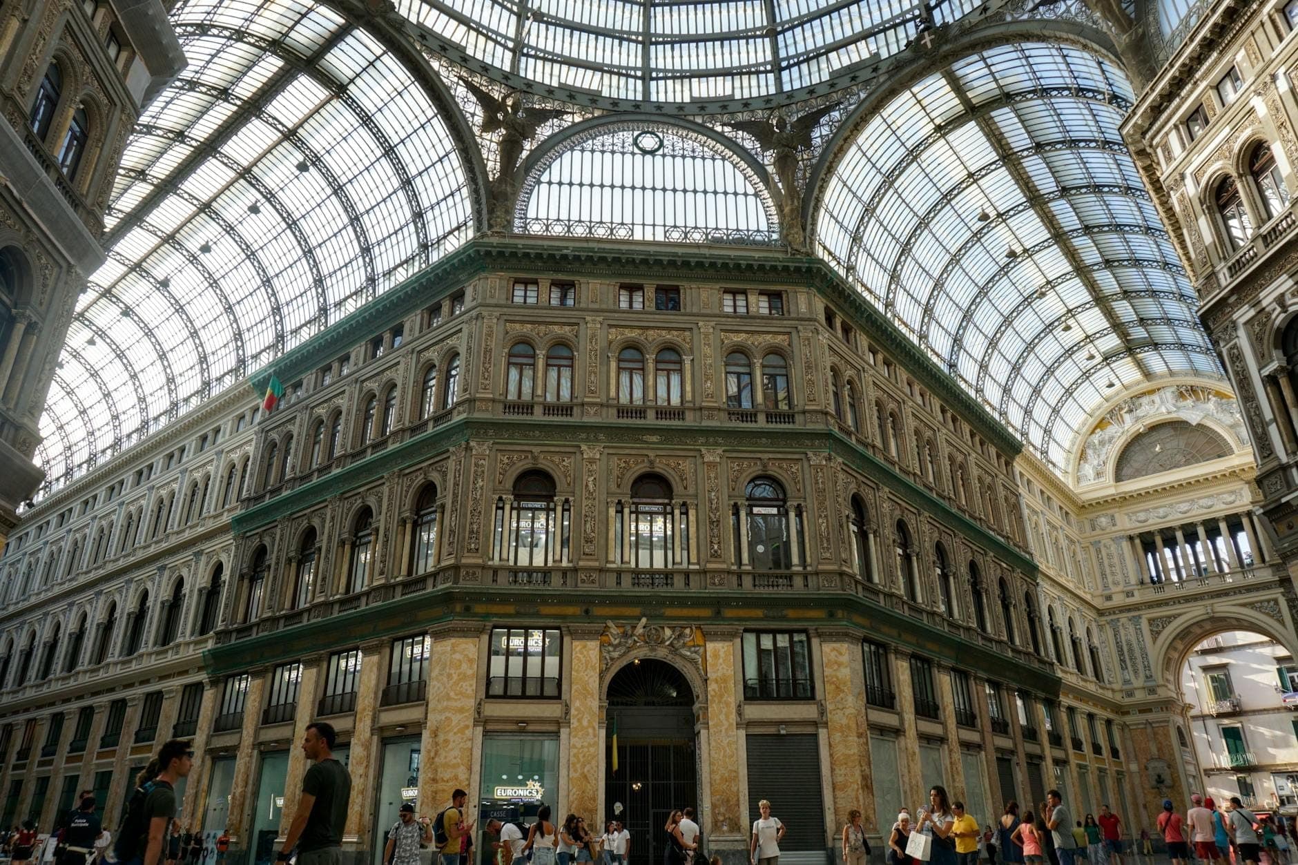 Pessoas caminhando e se reunindo sob a grande cúpula de vidro da Galleria Umberto I em Nápoles, com ornamentada arquitetura histórica.