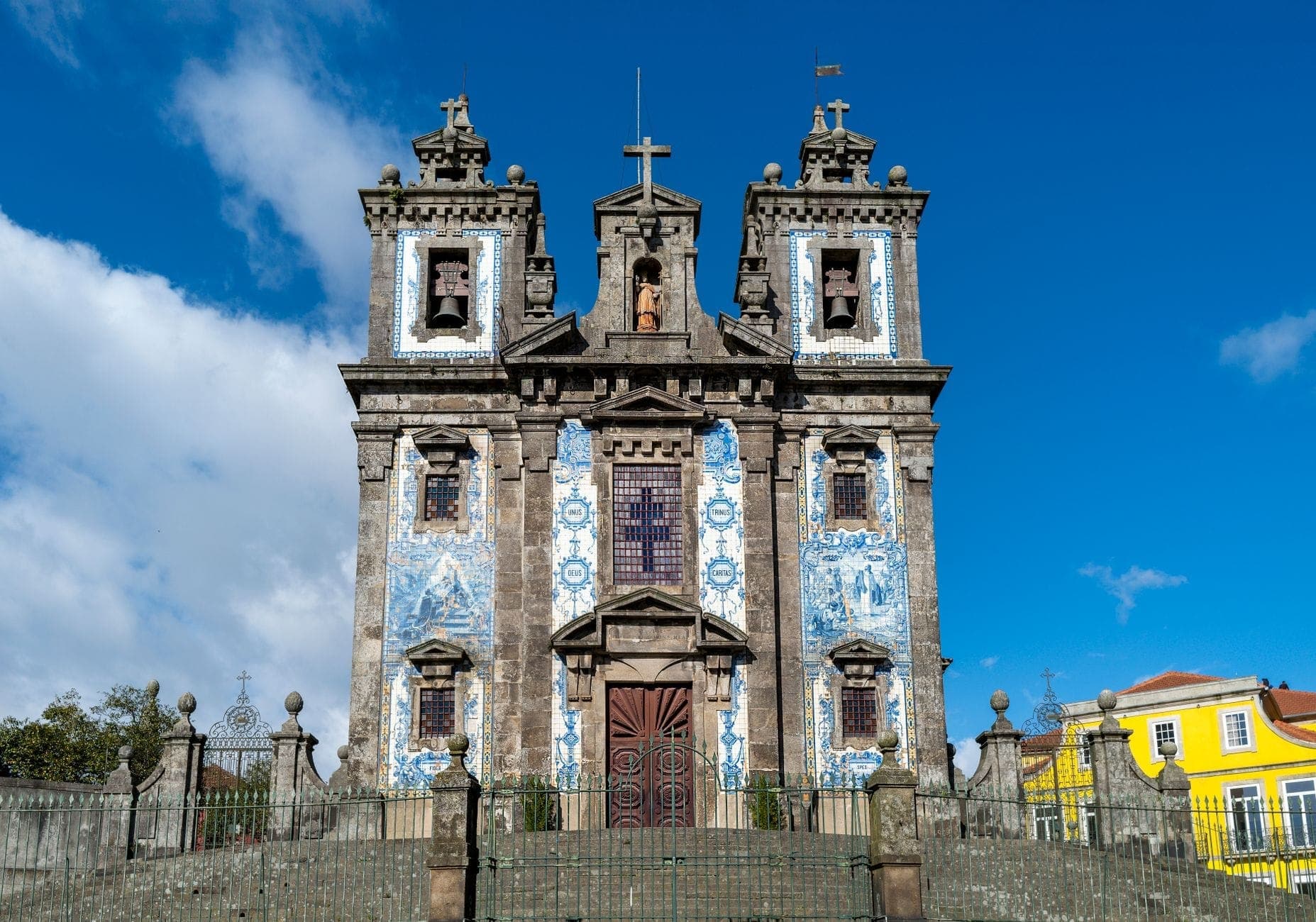 Frontansicht der Igreja de Santo Ildefonso in Porto mit der ikonischen blau-weißen Azulejo-Fassade unter strahlendem blauen Himmel und bunten Gebäuden in der Umgebung.