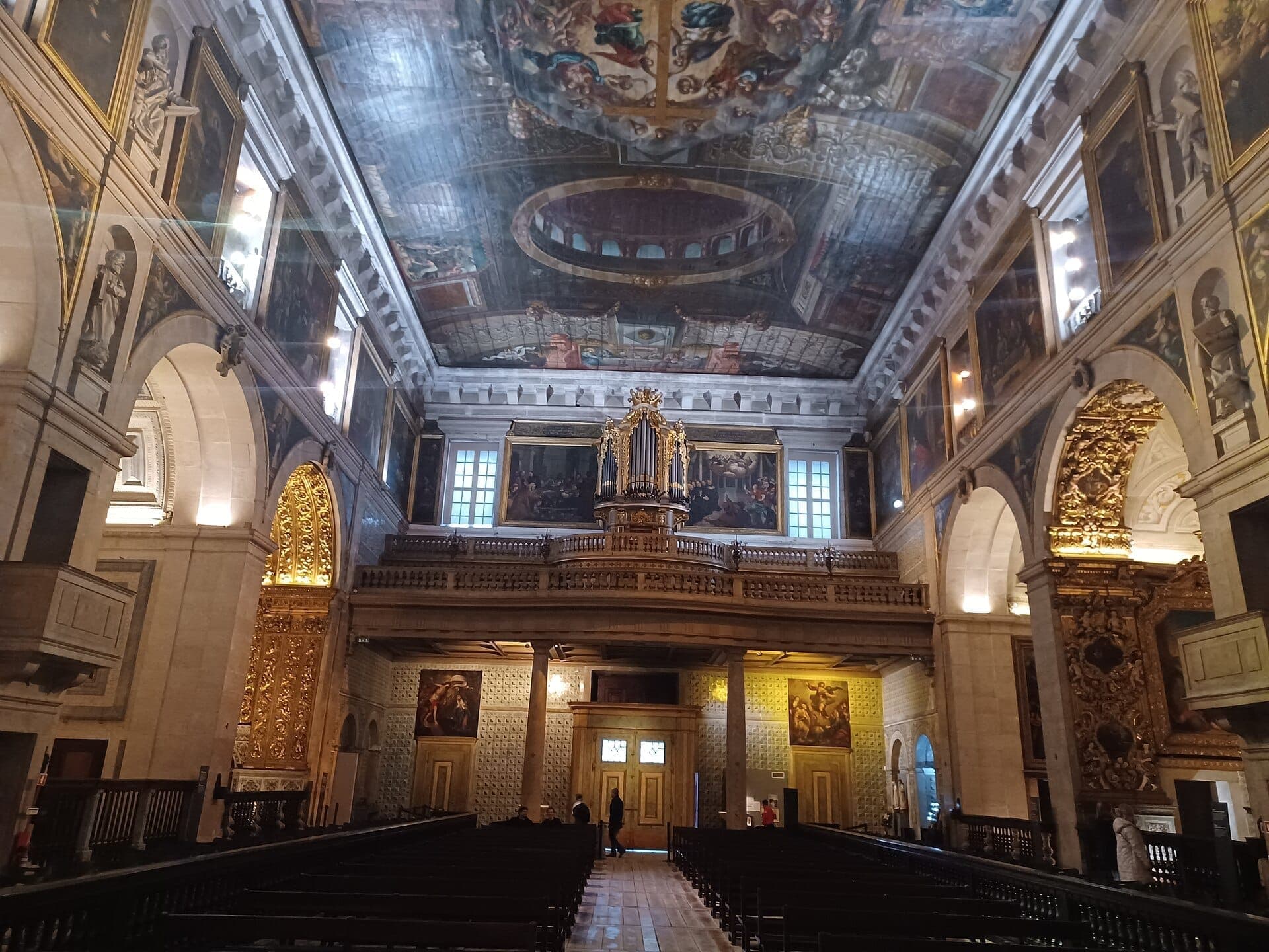 Vista interior da Igreja de São Roque com frescos intrincados no tecto, altares dourados, pinturas clássicas e bancos de madeira em luz natural suave.